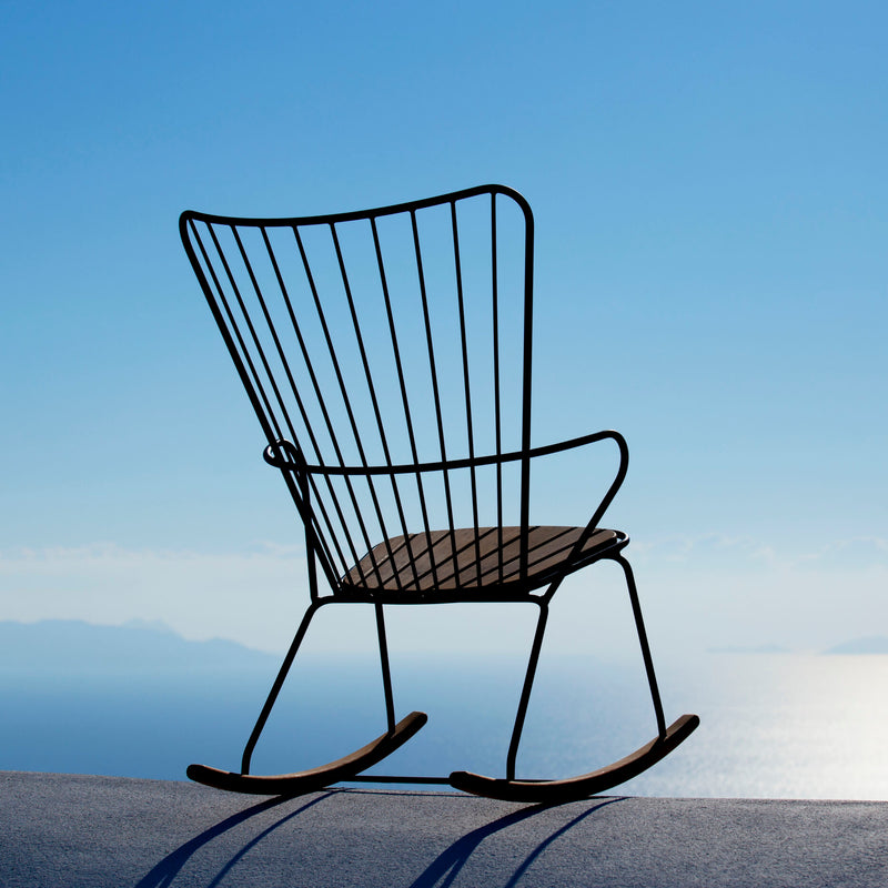 A Houe Paon Rocking Chair, crafted from powder-coated black steel, is set on concrete, offering a view of serene waters beneath a clear blue sky.