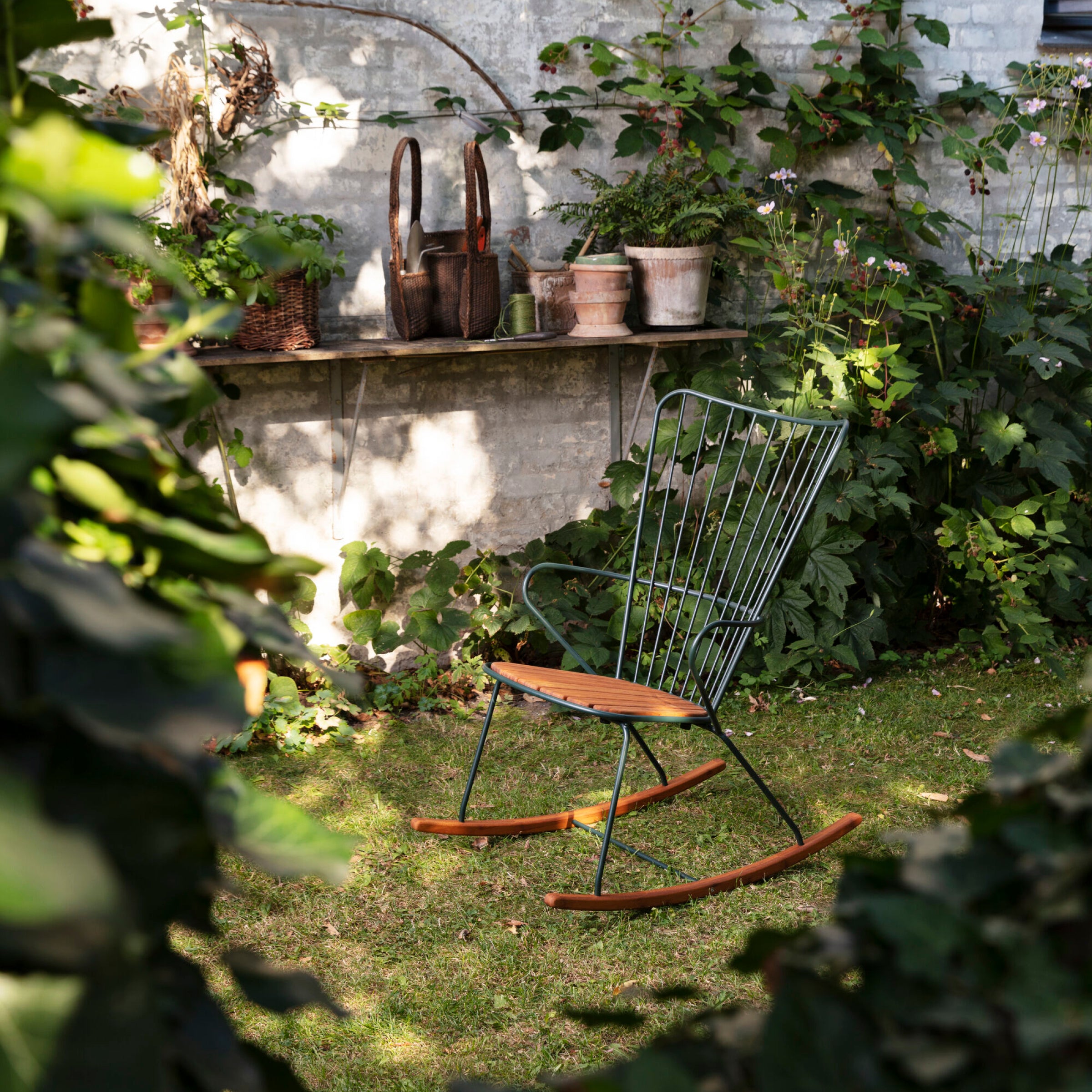 The Paon Rocking Chair by Houe, featuring green bamboo lamellas on metal, sits on a sunlit lawn. Behind it is a stone shelf on a brick wall with pots and baskets. Lush greenery and plants surround the area, creating a peaceful atmosphere.