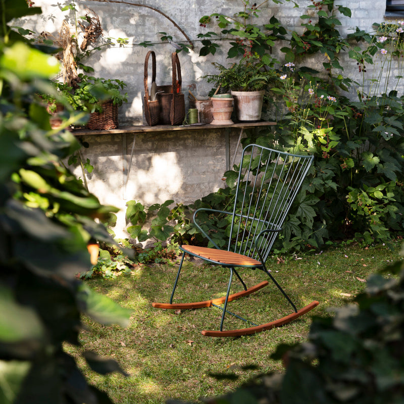The Paon Rocking Chair by Houe, featuring green bamboo lamellas on metal, sits on a sunlit lawn. Behind it is a stone shelf on a brick wall with pots and baskets. Lush greenery and plants surround the area, creating a peaceful atmosphere.