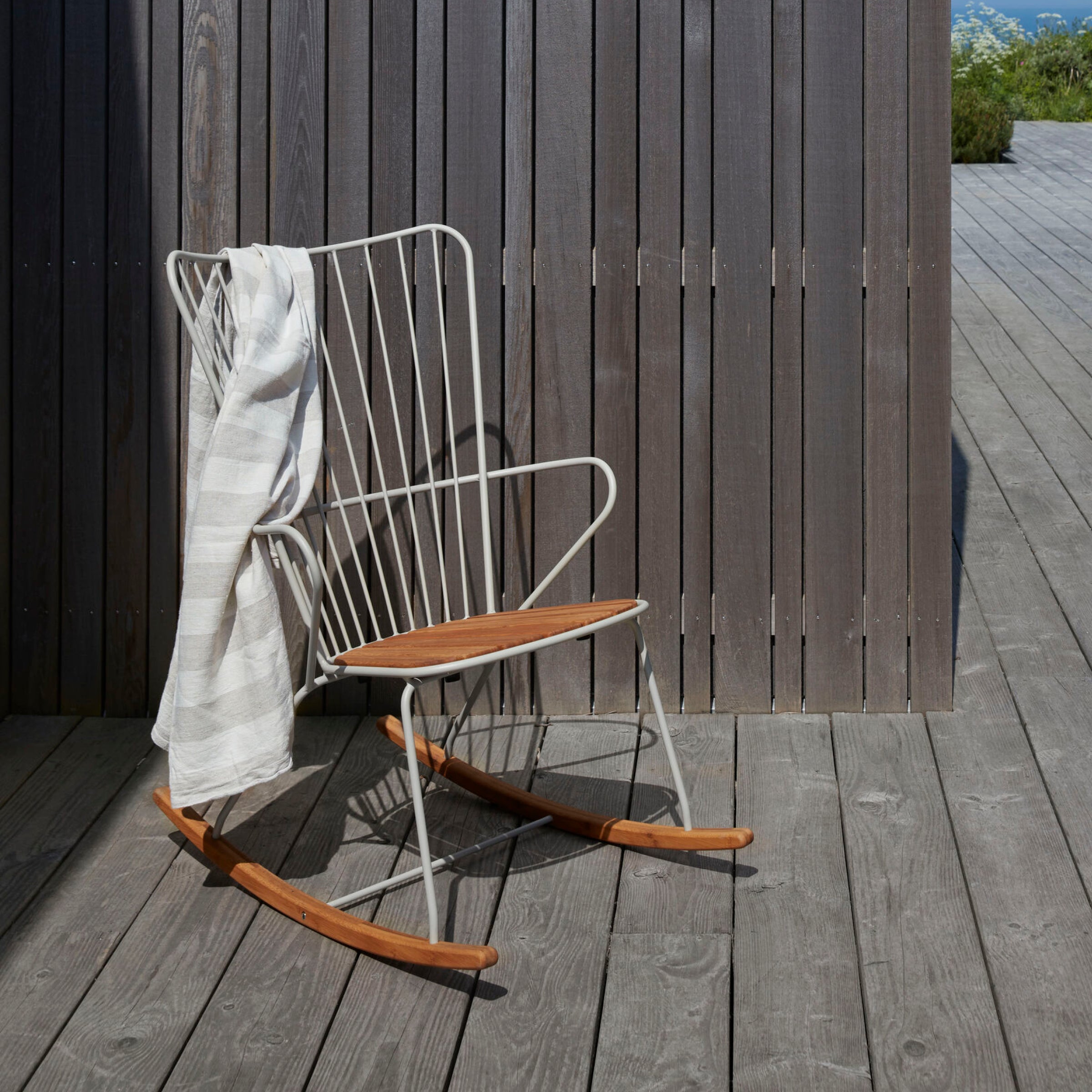 The Paon Rocking Chair by Houe, a blend of wood and powder-coated steel, is adorned with a light striped blanket. Its set on a wooden deck against bamboo lamellas, with white flowers and green foliage in the background.