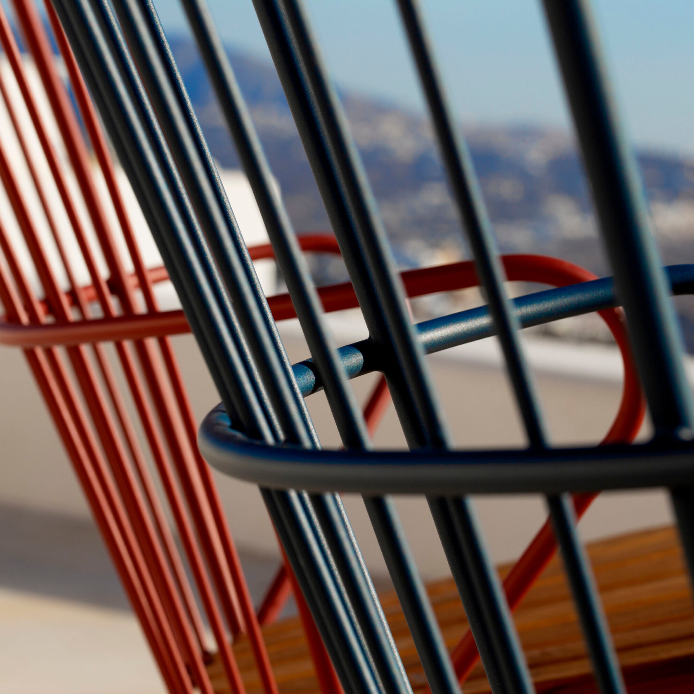 On a balcony, two Houe Paon Rocking Chairs with red and black powder-coated steel frames boast vertical bars, set against a blurred mountainous landscape and clear blue sky, contrasting beautifully with natures grandeur.