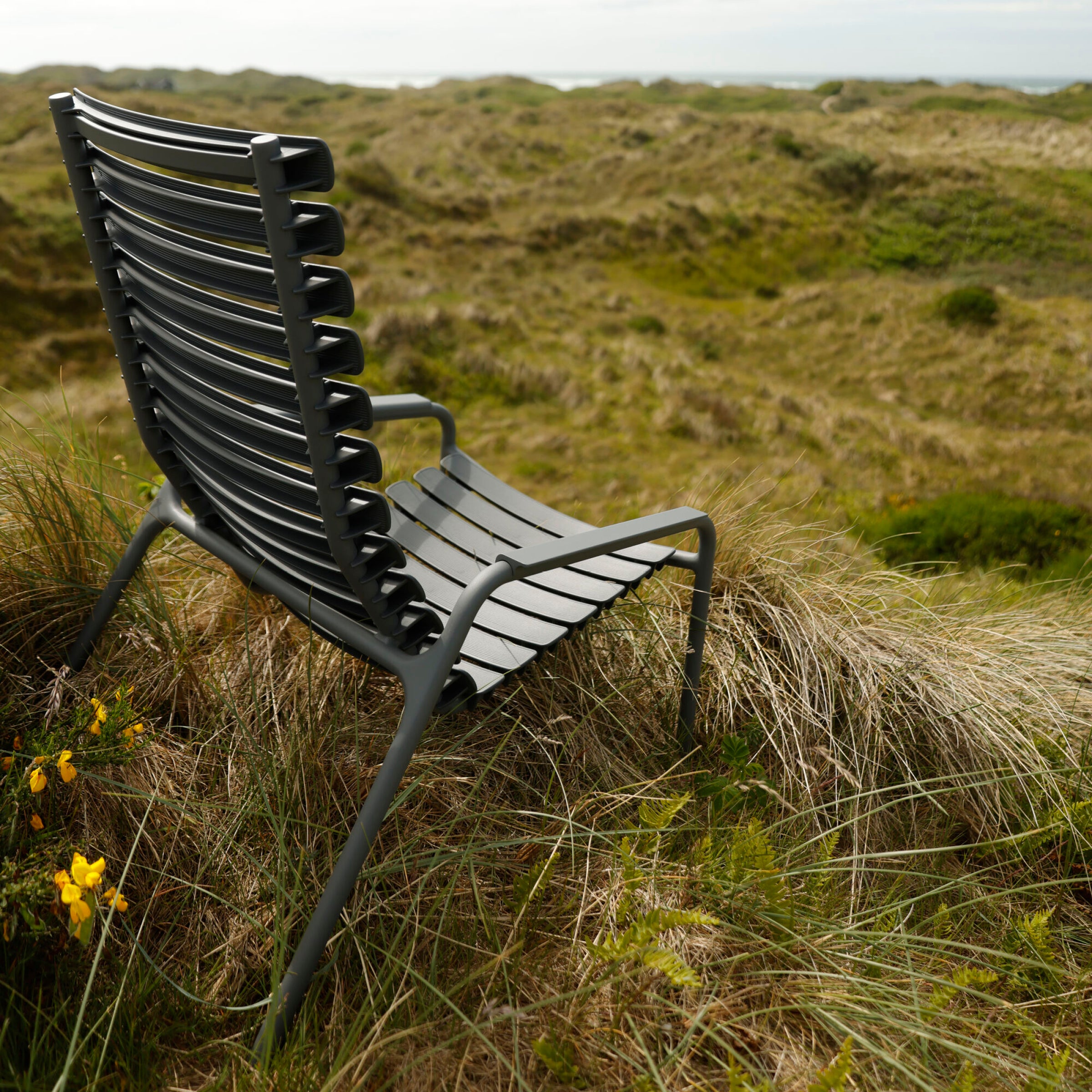 The ReClips Lounge Chair by Houe, inspired by Danish design, sits on grassy dunes with rolling hills in the background and vibrant yellow flowers enhancing the scenes natural beauty beneath a cloudy sky.