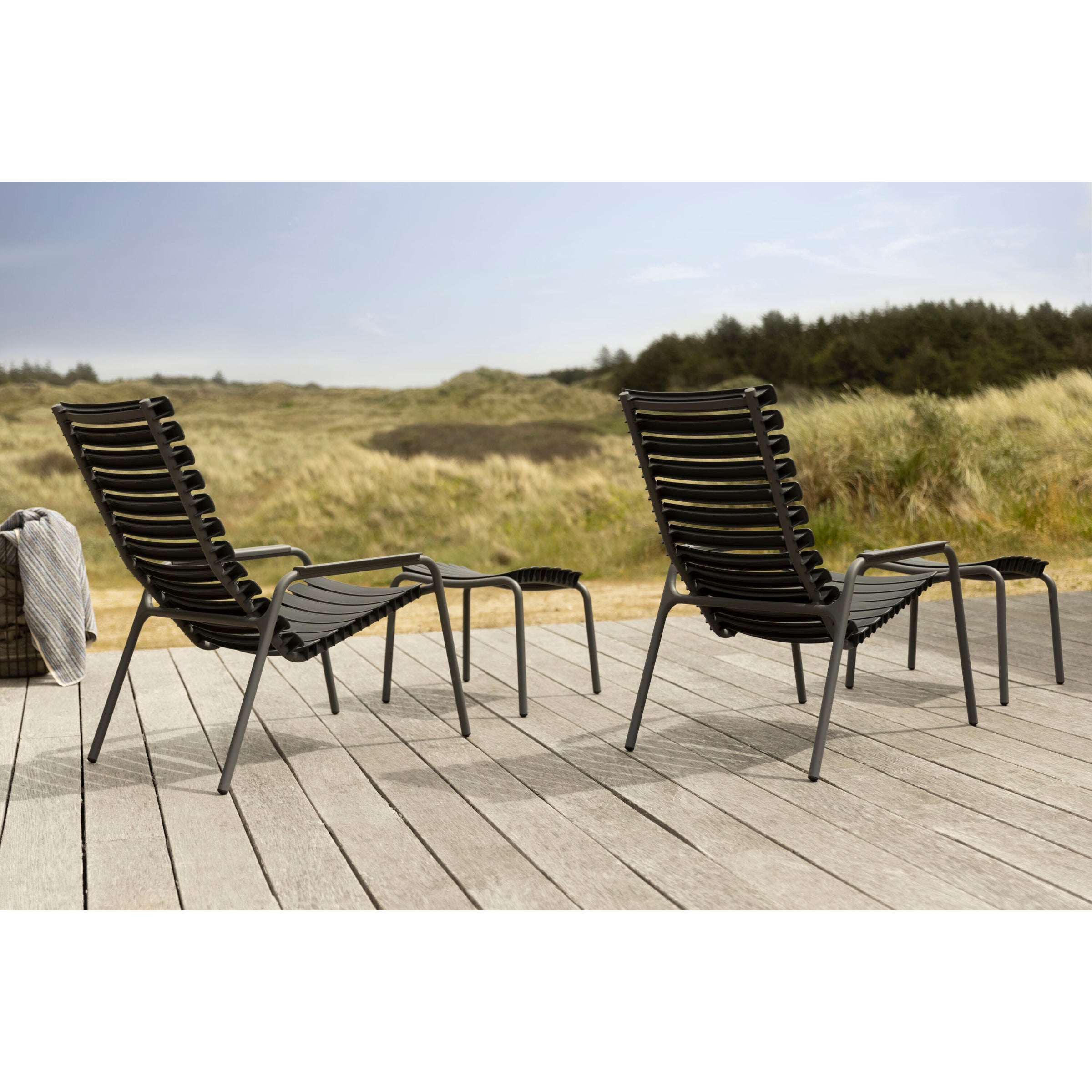 Two black Houe ReClips Lounge Chairs with footrests sit on a wooden deck facing grassy dunes under a clear sky. A folded striped towel rests nearby, highlighting the elegance of Danish design made from recycled plastic.