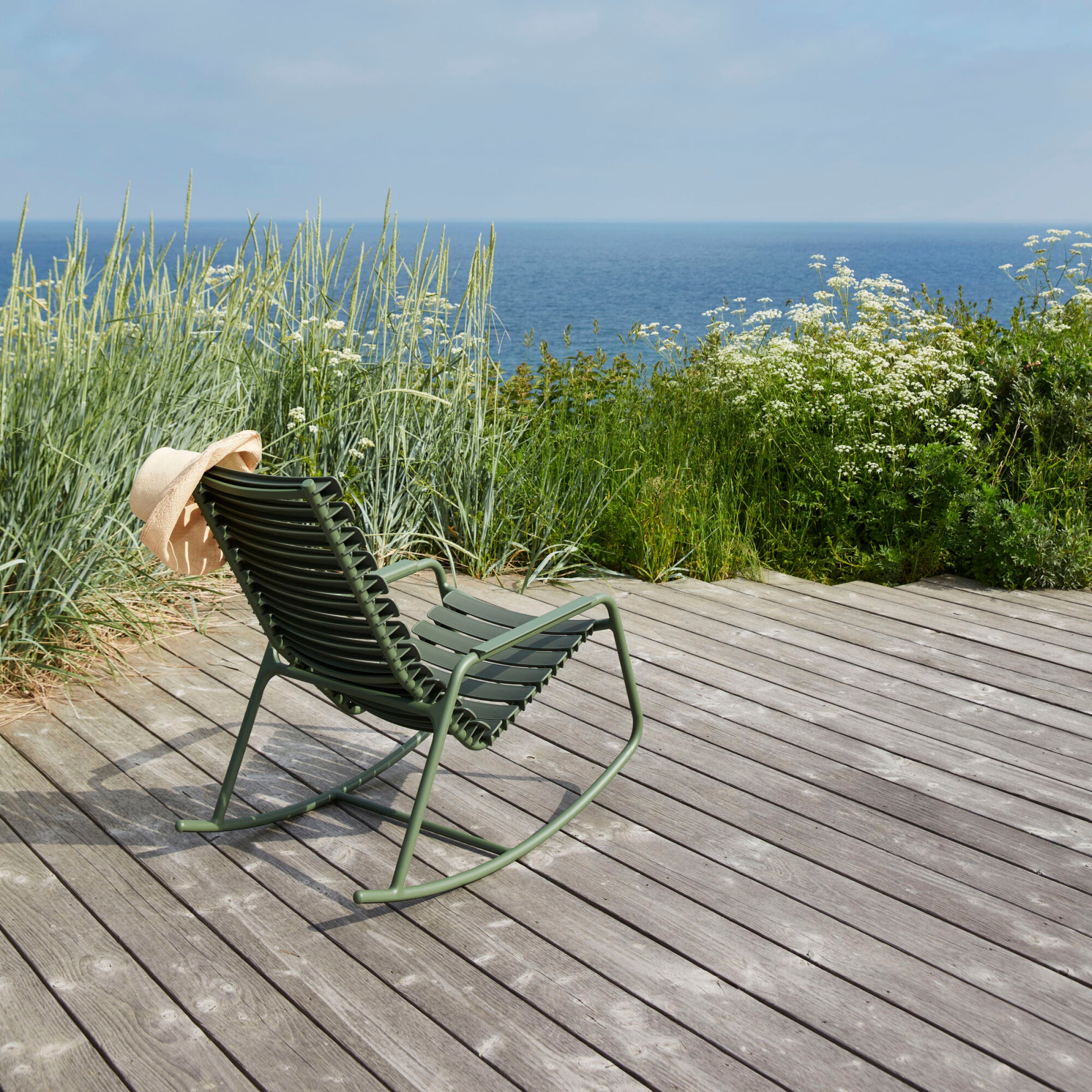 A Houe ReClips Rocking Chair with bamboo armrest and recycled green plastic lamellas sits on a wooden deck amid tall grass and wildflowers, while the blue ocean extends to the horizon under a cloudy sky. A light brown hat is casually placed on the chair.