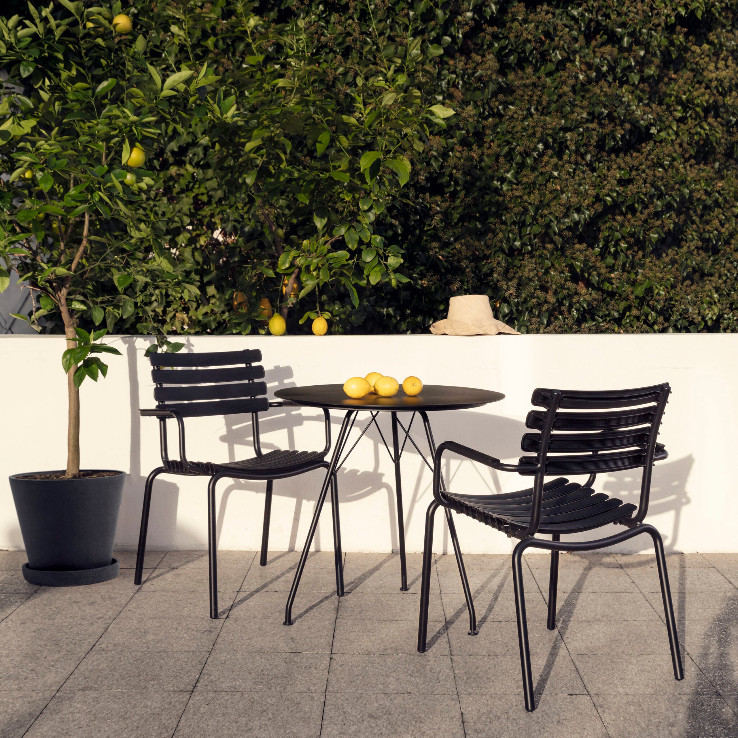 The outdoor scene features Houes ReClips Dining Chairs with sleek black metal frames showcasing Danish design. A round table holds lemons next to a thriving lemon tree, while a sun hat hangs on the wall surrounded by greenery—a serene spot embracing sustainability through recycled plastic.