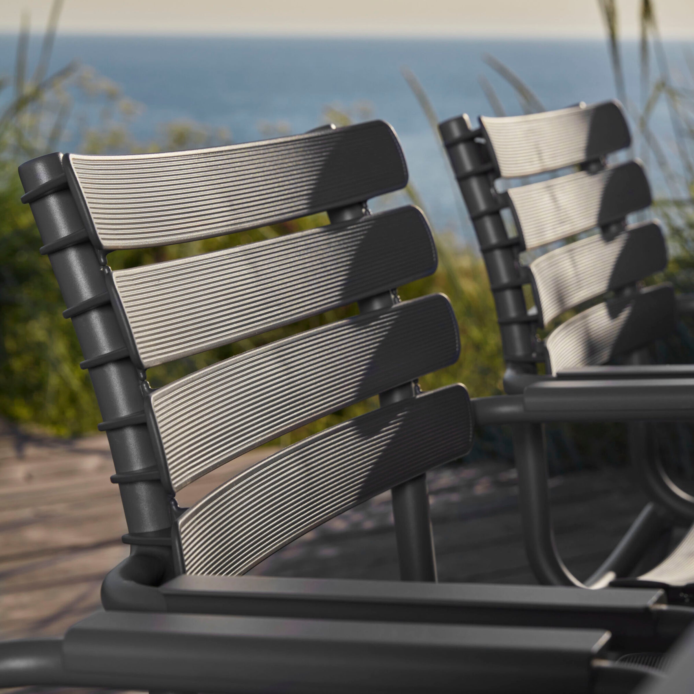 A pair of ReClips Dining Chairs by Houe, featuring sleek metal slats and black frames, sit on a wooden deck with a blurred backdrop of grassy plants and water. These chairs blend Danish design elegance with eco-friendly innovation using recycled plastic trash.