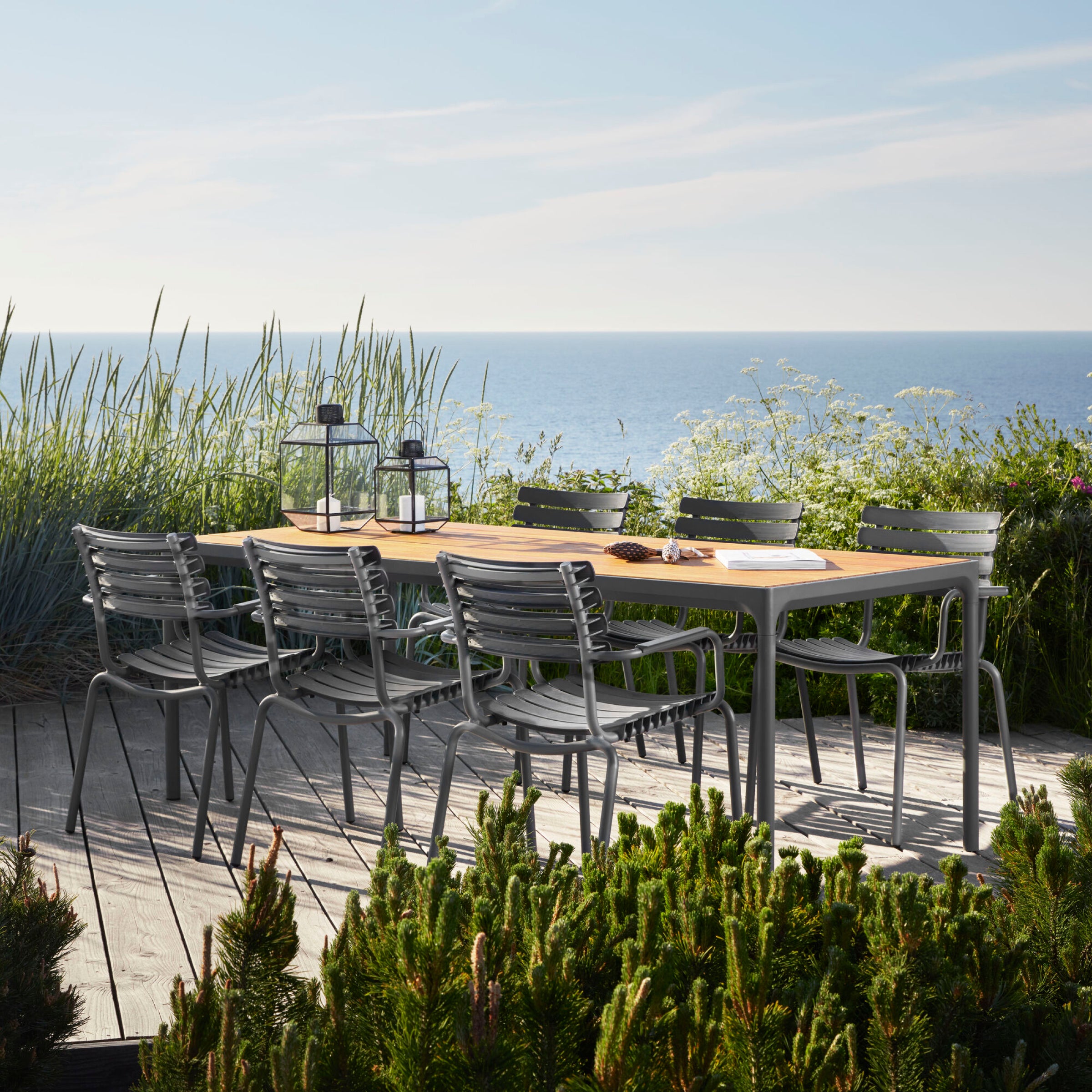 A wooden outdoor dining table with eight Houe ReClips Dining Chairs, showcasing Danish design, sits on a deck. Lanterns adorn it, surrounded by tall grass and wildflowers with an ocean view under a partly cloudy sky.