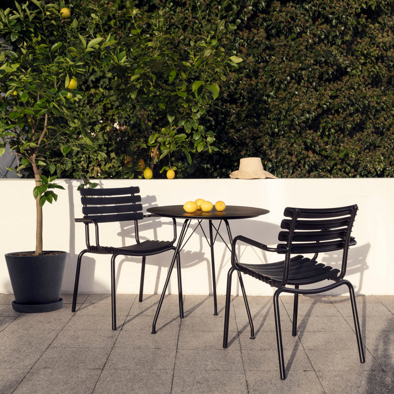 The outdoor scene features Houes ReClips Dining Chairs with sleek black metal frames showcasing Danish design. A round table holds lemons next to a thriving lemon tree, while a sun hat hangs on the wall surrounded by greenery—a serene spot embracing sustainability through recycled plastic.