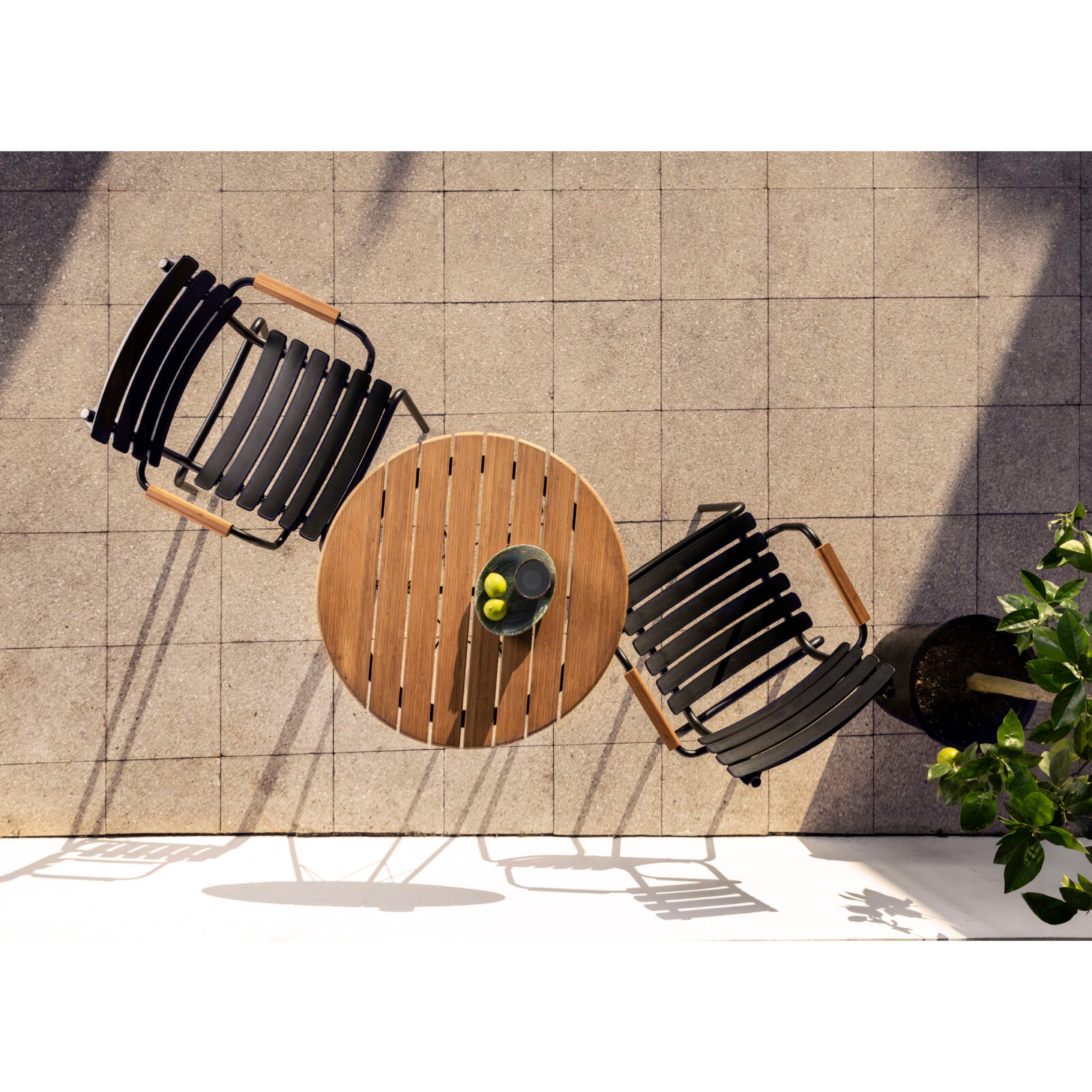 Aerial shot of a patio with Danish design: a round wooden table and two Houe ReClips Dining Chairs with Bamboo Armrests on tiles. The table holds a sleek black pitcher, two lemons, surrounded by shadows and greenery.