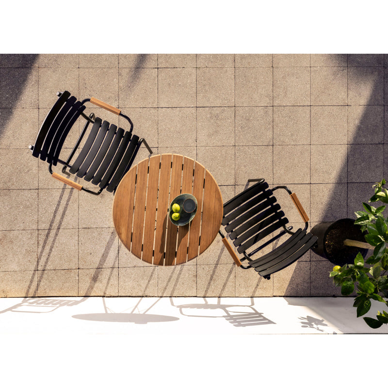 Aerial shot of a patio with Danish design: a round wooden table and two Houe ReClips Dining Chairs with Bamboo Armrests on tiles. The table holds a sleek black pitcher, two lemons, surrounded by shadows and greenery.