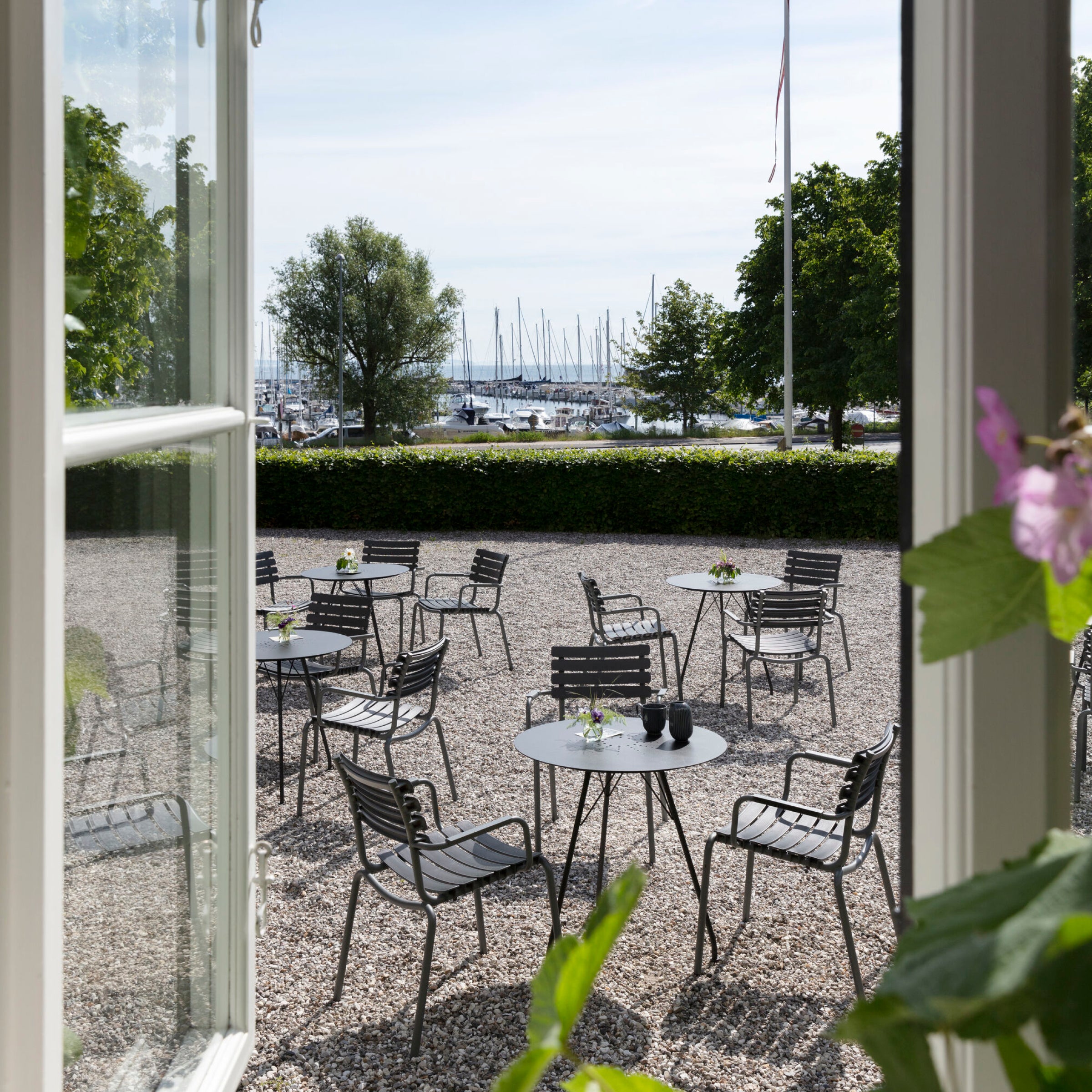 Relax in an outdoor setting with ReClips Dining Chairs by Houe, crafted from recycled plastic. Surrounded by lush trees and a gravel surface, enjoy views of sailboats swaying at the marina under a clear sky, enhancing the Danish designs serene charm.