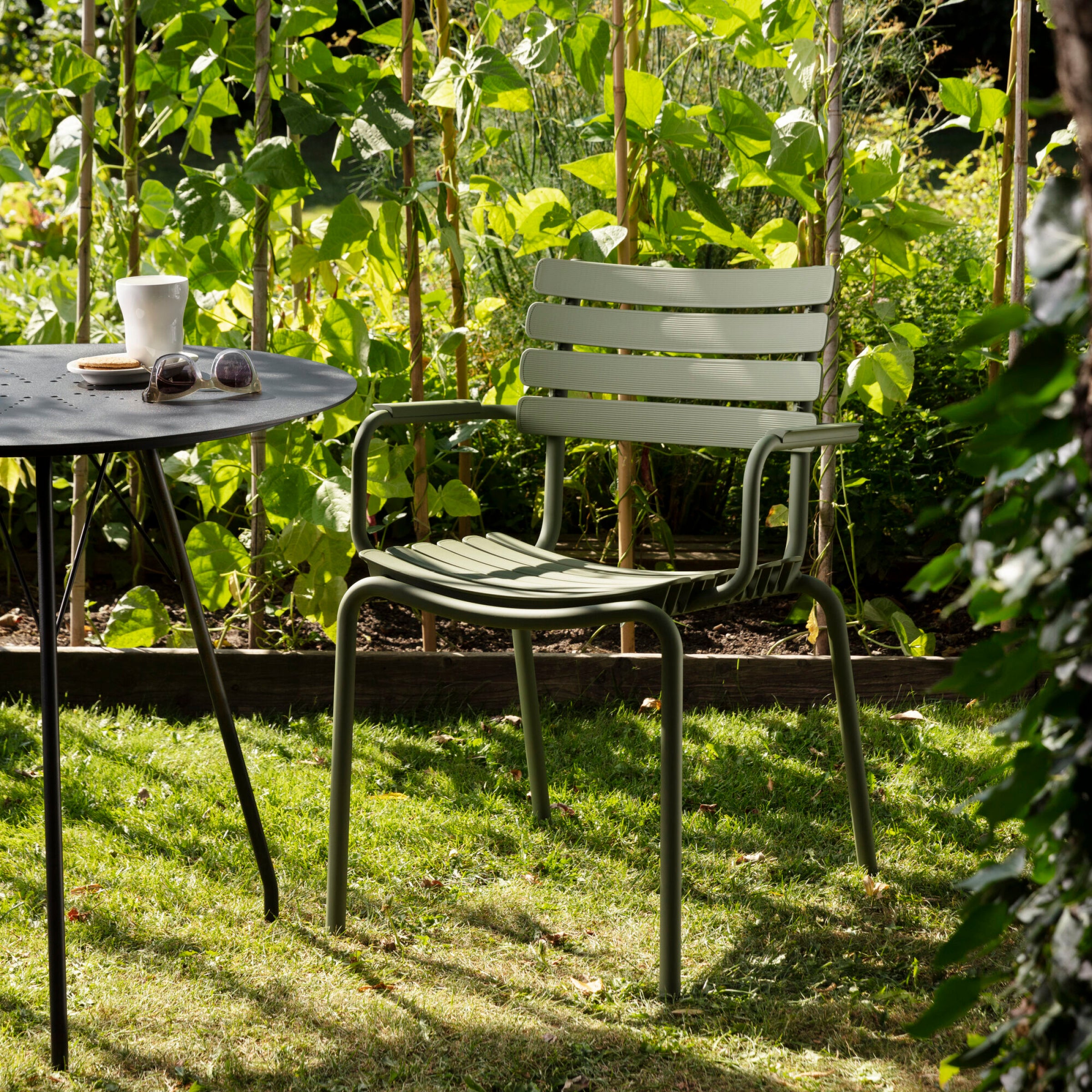 A ReClips Dining Chair from Houe, in green metal, is set on garden grass reflecting modern Danish style. Nearby, a round black table holds sunglasses, a coffee cup, and a cookie while sunlight streams through the tall greenery.