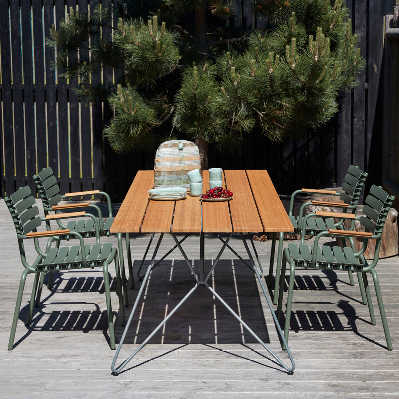 The ReClips Dining Chair with Bamboo Armrest by Houe complements the outdoor wooden table on a deck. Teal ceramic dishes, cups, a vase, and a small bowl of cherries add inviting touches. A pine tree and dark wooden fence frame the Danish-designed scene.
