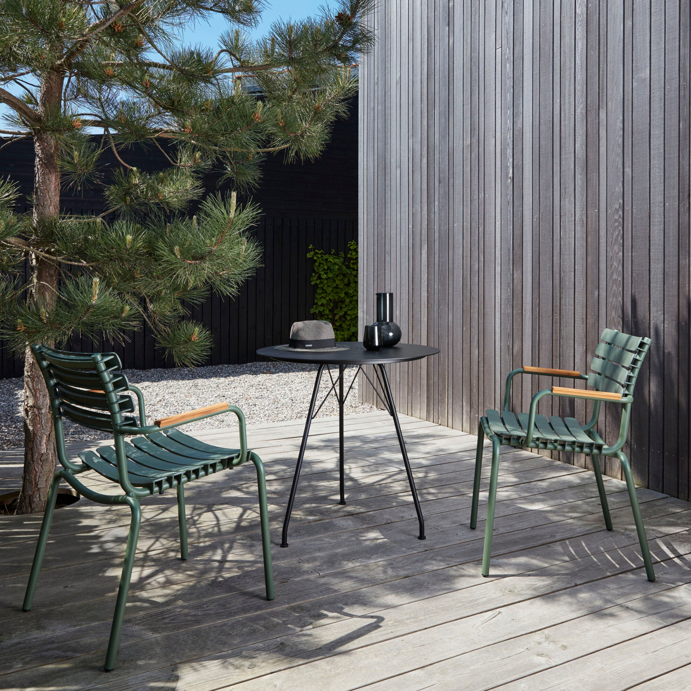 A minimalist patio showcases Danish design with two green Houe ReClips Dining Chairs featuring plastic lamellas and bamboo armrests, alongside a round black table with vases. This setup sits on a wooden deck near pine and gravel, framed by vertical wood paneling.