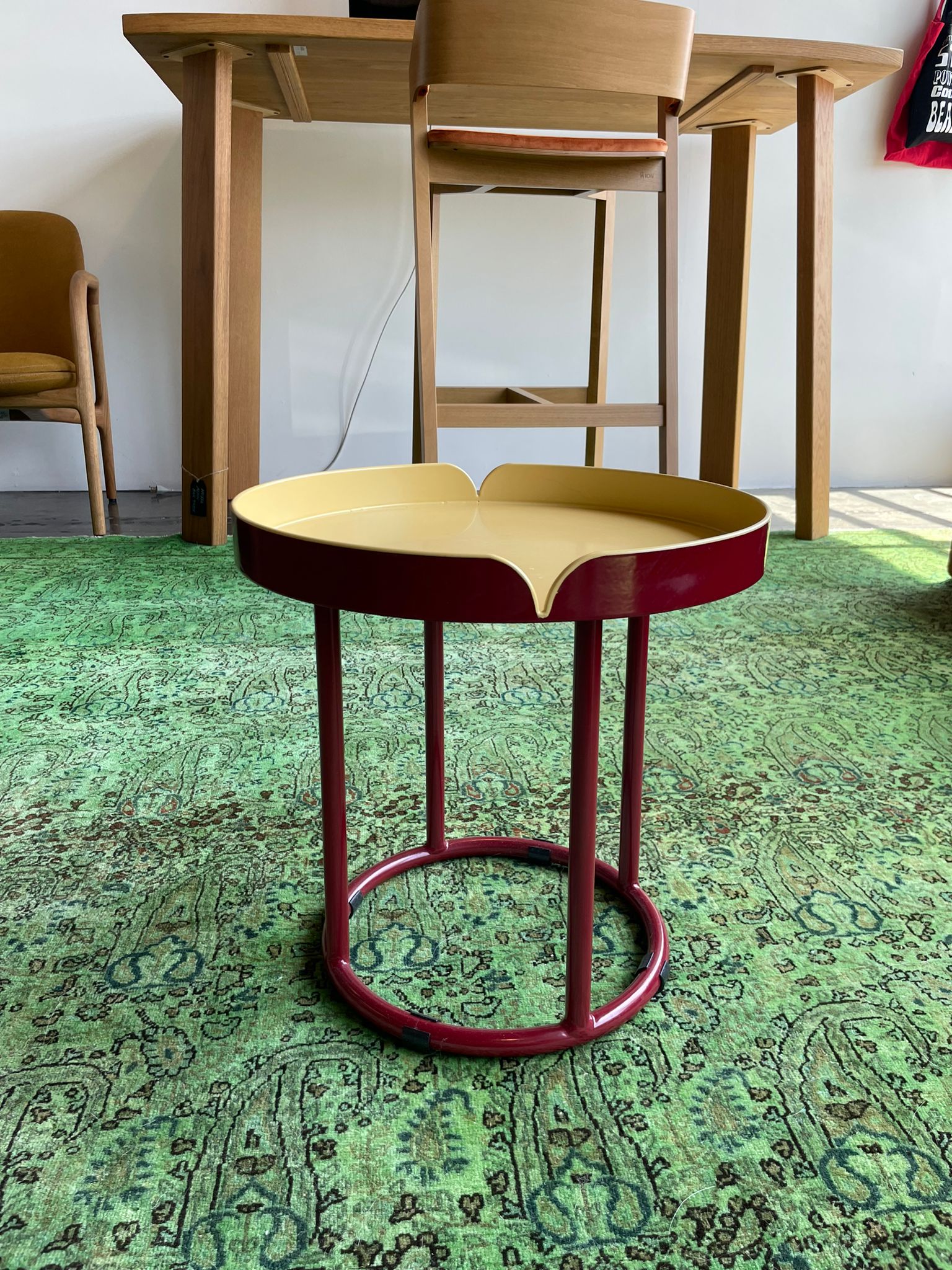 A small, round table from Ames Victorias Coffee Table - Inventory series has a red base and yellow scalloped-edge top, placed on a green patterned rug. Wooden chairs and another table are in the background of this bright, naturally lit room.