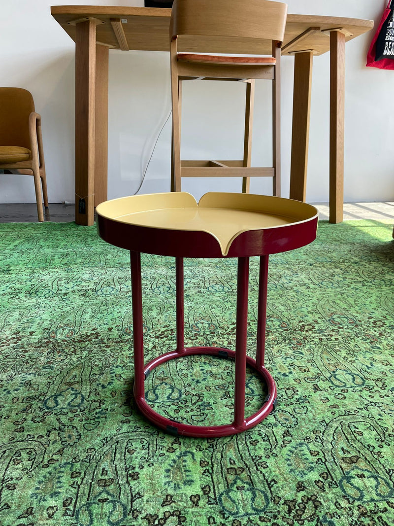 A small, round table from Ames Victorias Coffee Table - Inventory series has a red base and yellow scalloped-edge top, placed on a green patterned rug. Wooden chairs and another table are in the background of this bright, naturally lit room.