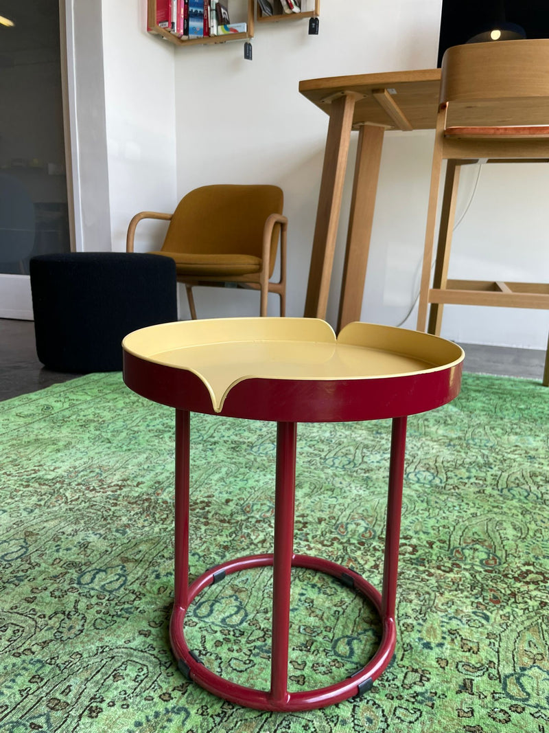 A Victorias Coffee Table from Ames, featuring a red base and yellow top, sits on a patterned green rug. In the background is a wooden chair with a mustard cushion, a black ottoman, and part of an Ames Designer Furniture desk. Wall shelves filled with books complete the scene.