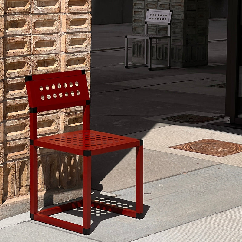 Red metal chair on a sidewalk with textured wall and another chair in the background.