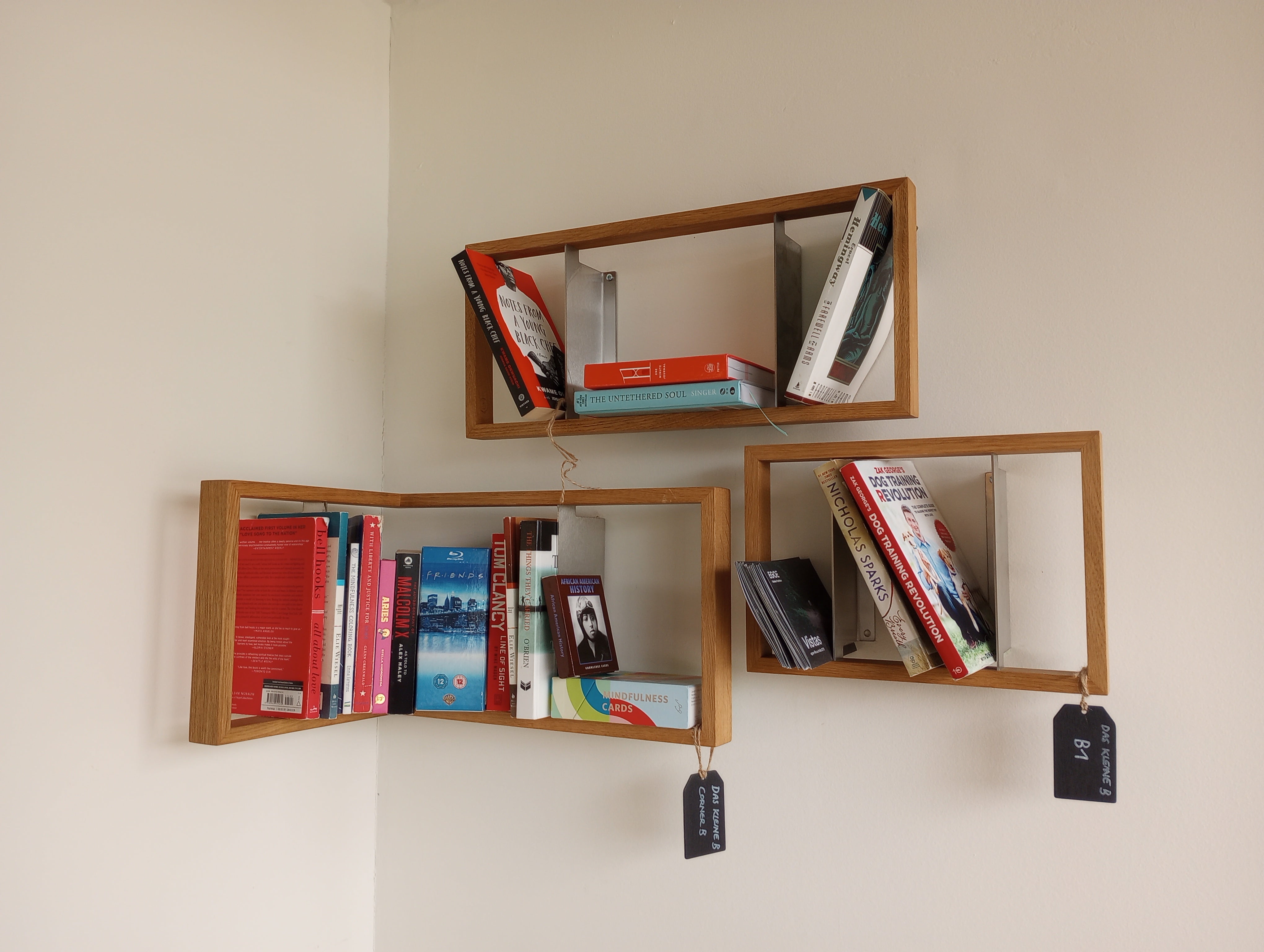 Three b 8.7 - Sale bookshelves by Das kleine b, with solid wooden frames, are mounted on a white corner wall. Each shelf holds colorful books with small black tags dangling, creating a unique floating display.