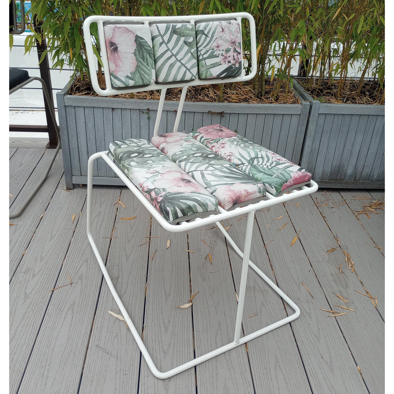 A comfortable Hope Chair by Egoe, featuring a tropical-themed cushion with green palm leaves and pink flowers, sits on a wooden deck. The background showcases tall planters with bamboo plants for an inviting oasis.