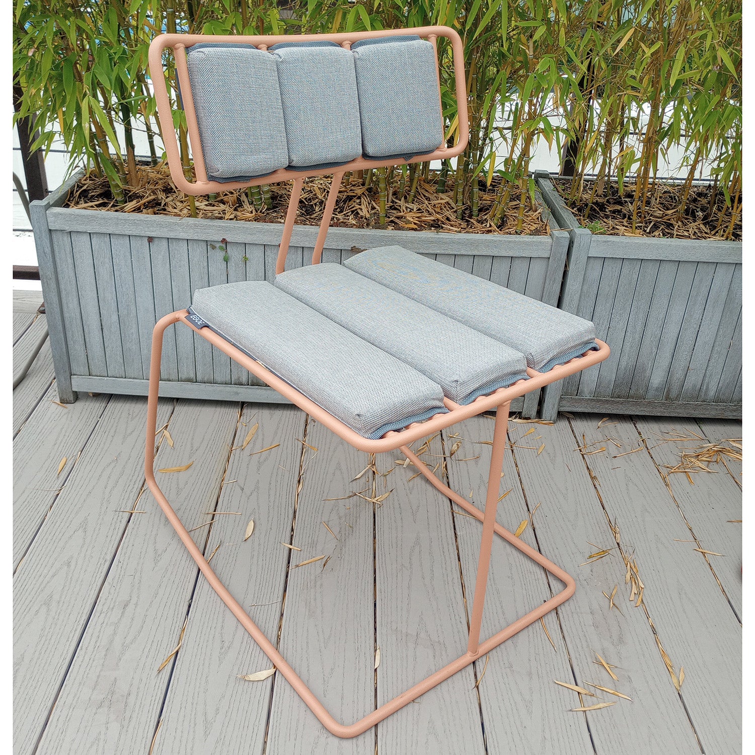 A Hope Chair - SALE by Egoe, with a pink steel frame and light gray padded cushions, graces the wooden deck. Adorned with three back and seat cushions, its framed by bamboo-filled planters, adding style and serenity to the outdoor setting.