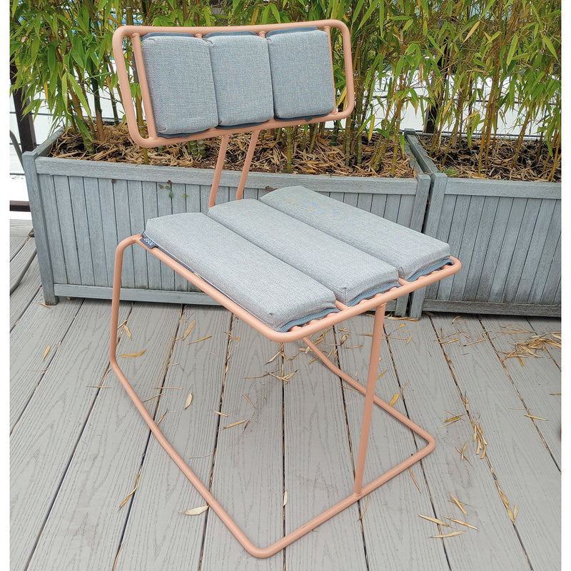 A Hope Chair - SALE by Egoe, with a pink steel frame and light gray padded cushions, graces the wooden deck. Adorned with three back and seat cushions, its framed by bamboo-filled planters, adding style and serenity to the outdoor setting.