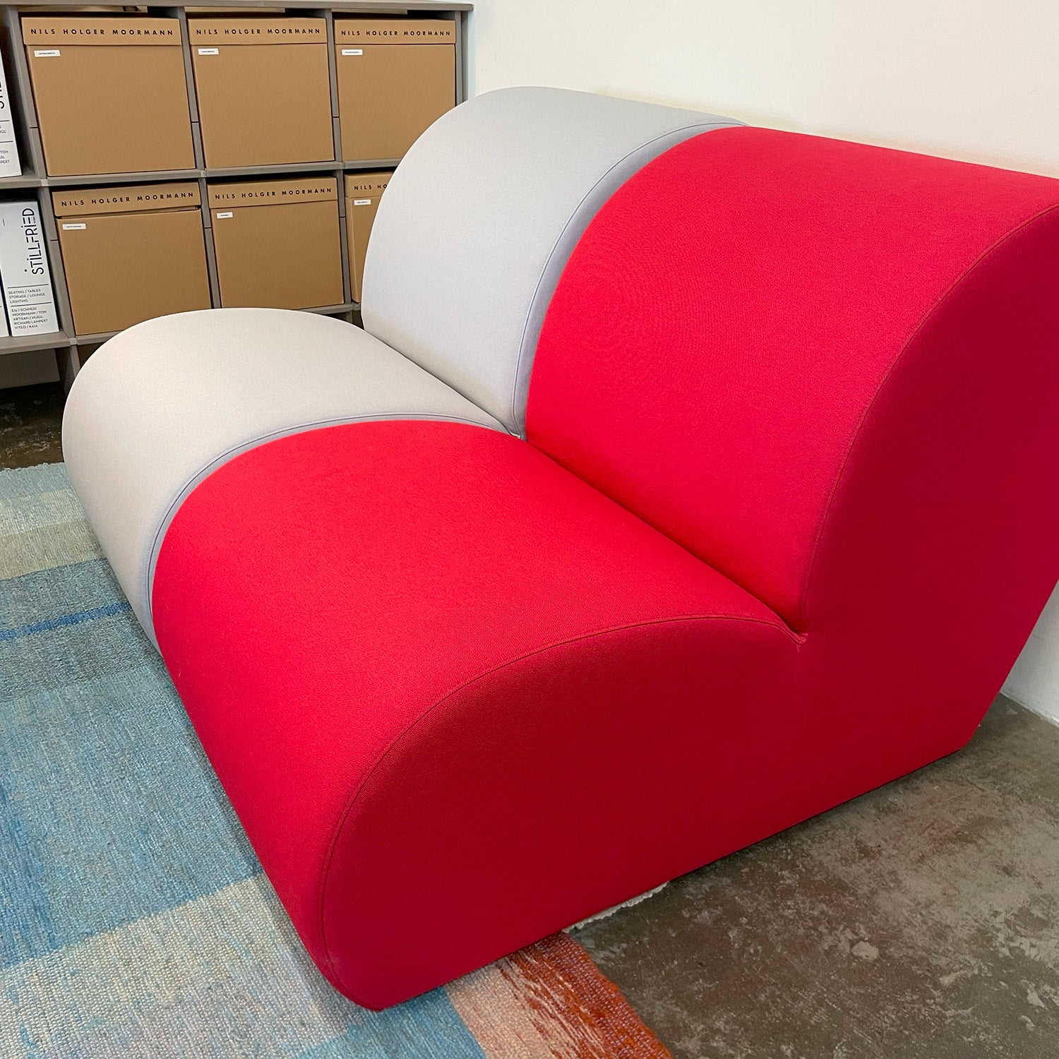 The Levi Seating Lounge Chair - SALE by Objekte unserer Tage highlights a bold red and light gray color-block design atop a multicolored rug, with shelves filled with brown cardboard storage boxes in the background creating an organized, minimalist ambiance.