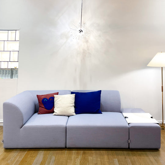 A modern living room features the Objekte unserer Tage Weber Modular Sofa in light gray. Adorned with three pillows—red with a blue heart, white, and blue—and an attached sleek white coffee table, a decorative chandelier elegantly hangs from above.