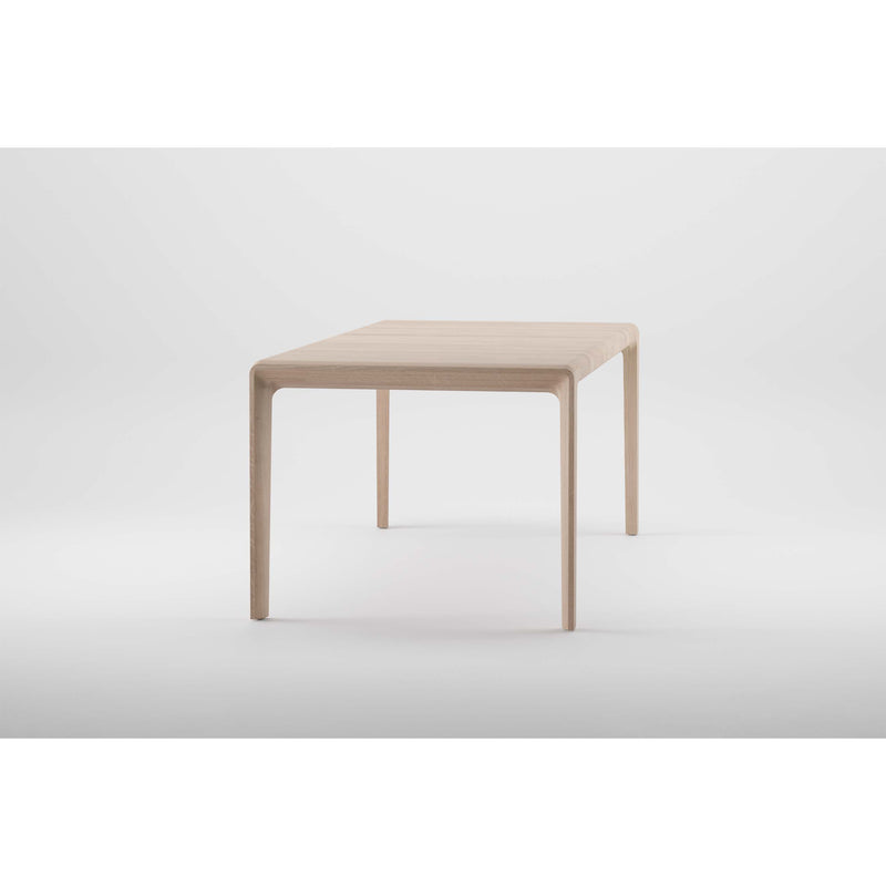 The Artisan INVITO Table is a minimalist, light wood table made from solid, eco-friendly materials. It has a smooth, rectangular top supported by four slender legs and is showcased against a plain light gray background.