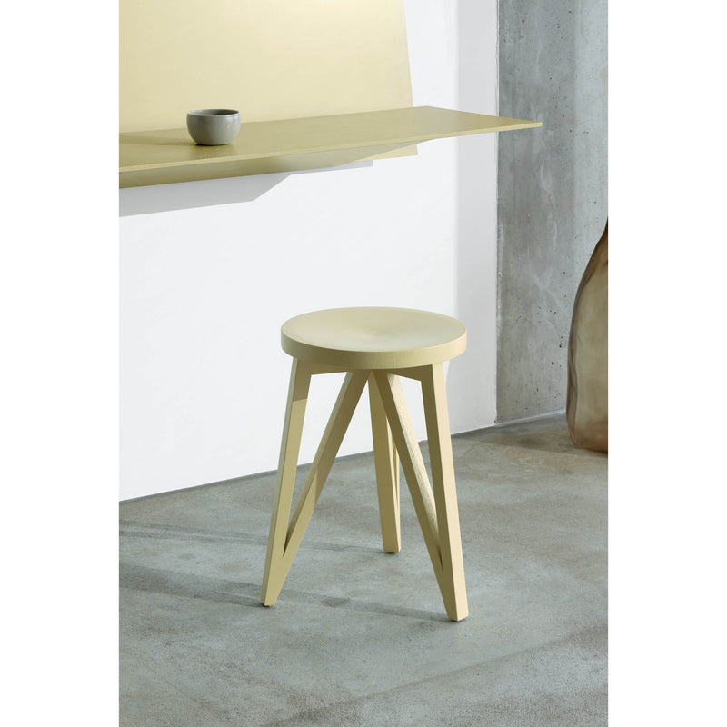 A minimalist JL1 Faber Stool by Loehr with a round seat and triangular legs stands gracefully on a gray concrete floor. In the background, a matching desk from the FABER collection showcases northern European design, elegantly holding a small ceramic bowl.