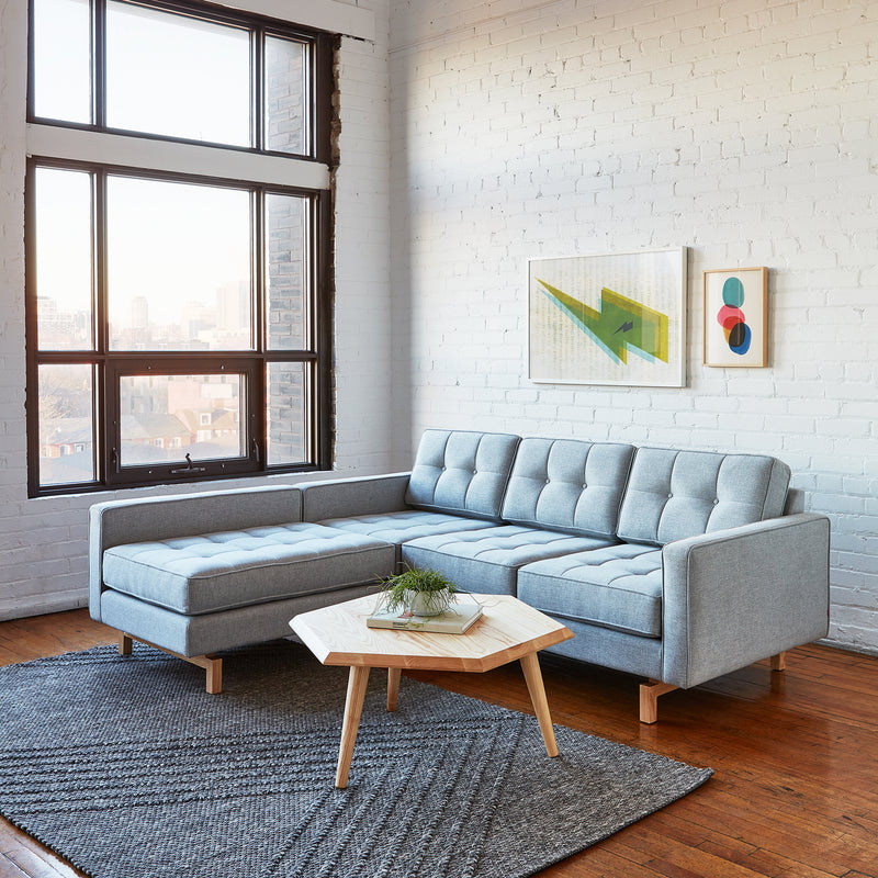 A modern living room featuring a light gray Jane 2 LOFT Bi-Sectional Natural Finish Base by Gus Modern, a wooden hexagonal coffee table with a small plant, dark textured rug, large windows, white brick walls, and two colorful abstract artworks.