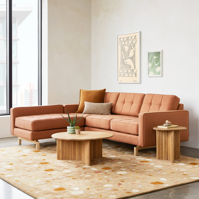 A modern living room featuring a Gus Modern Jane 2 LOFT Bi-Sectional with a natural finish base in burnt orange, two round wooden coffee tables, a patterned rug, pillows, potted plants, and wall art by a large window.