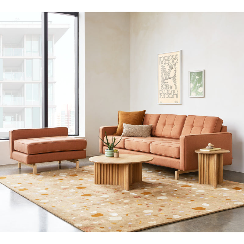 A modern living room featuring a Gus Modern Jane 2 LOFT Bi-Sectional with a natural finish base in orange, matching ottoman, two wooden tables, a beige patterned rug, wall art, and large windows framing breathtaking city views.