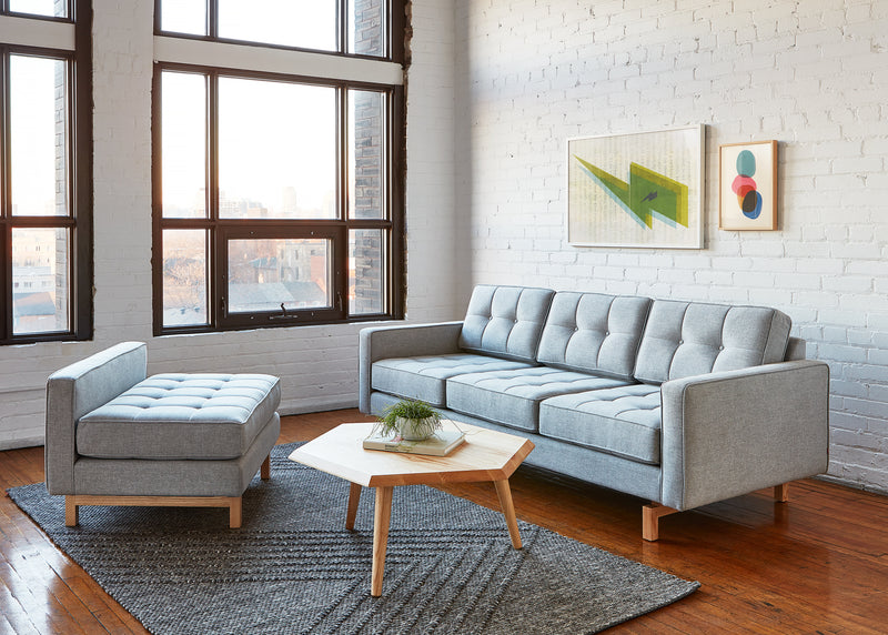 A modern living room featuring the Gus Modern Jane 2 LOFT Bi-Sectional with a natural finish base, paired with a matching ottoman, hexagonal coffee table, textured rug, wall art, and large windows highlighting white brick walls and wood floors.
