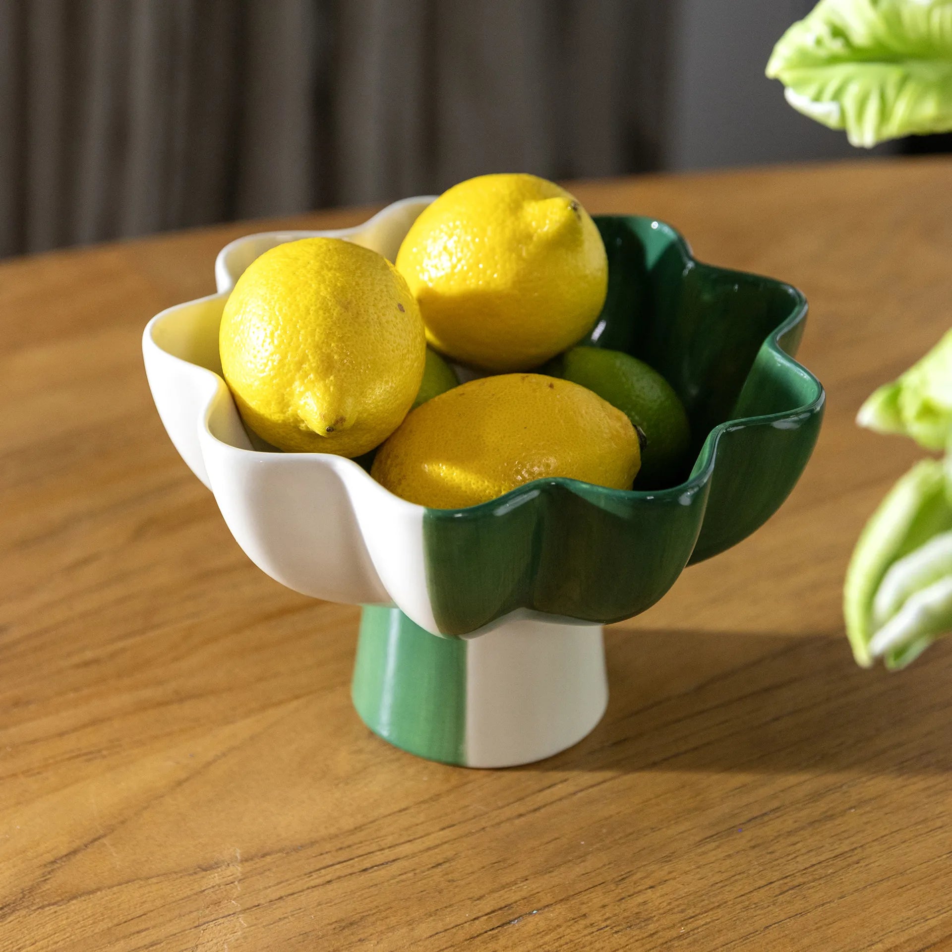 The &Klevering Fruit Bowl Sun Green Small, a multicolor ceramic piece with wavy edges and partly green and white design, artfully holds yellow lemons and a lime. It rests elegantly on a wooden table, framed by vibrant green leafy plants in the background.