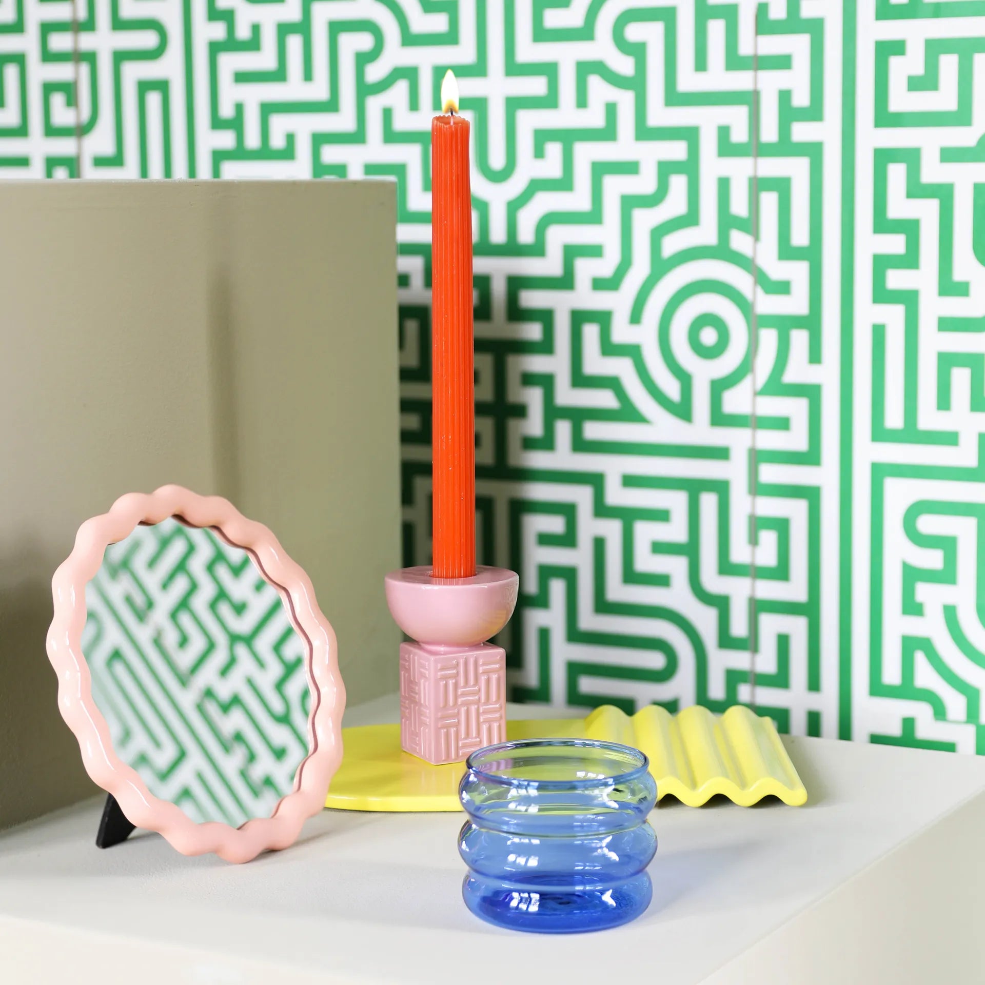 A decorative setup includes a &Klevering Candle Holder Stack Pink, alongside a pink wavy mirror, lit orange candle, yellow wavy tray, and a blue glass jar against a green and white geometric patterned wall.
