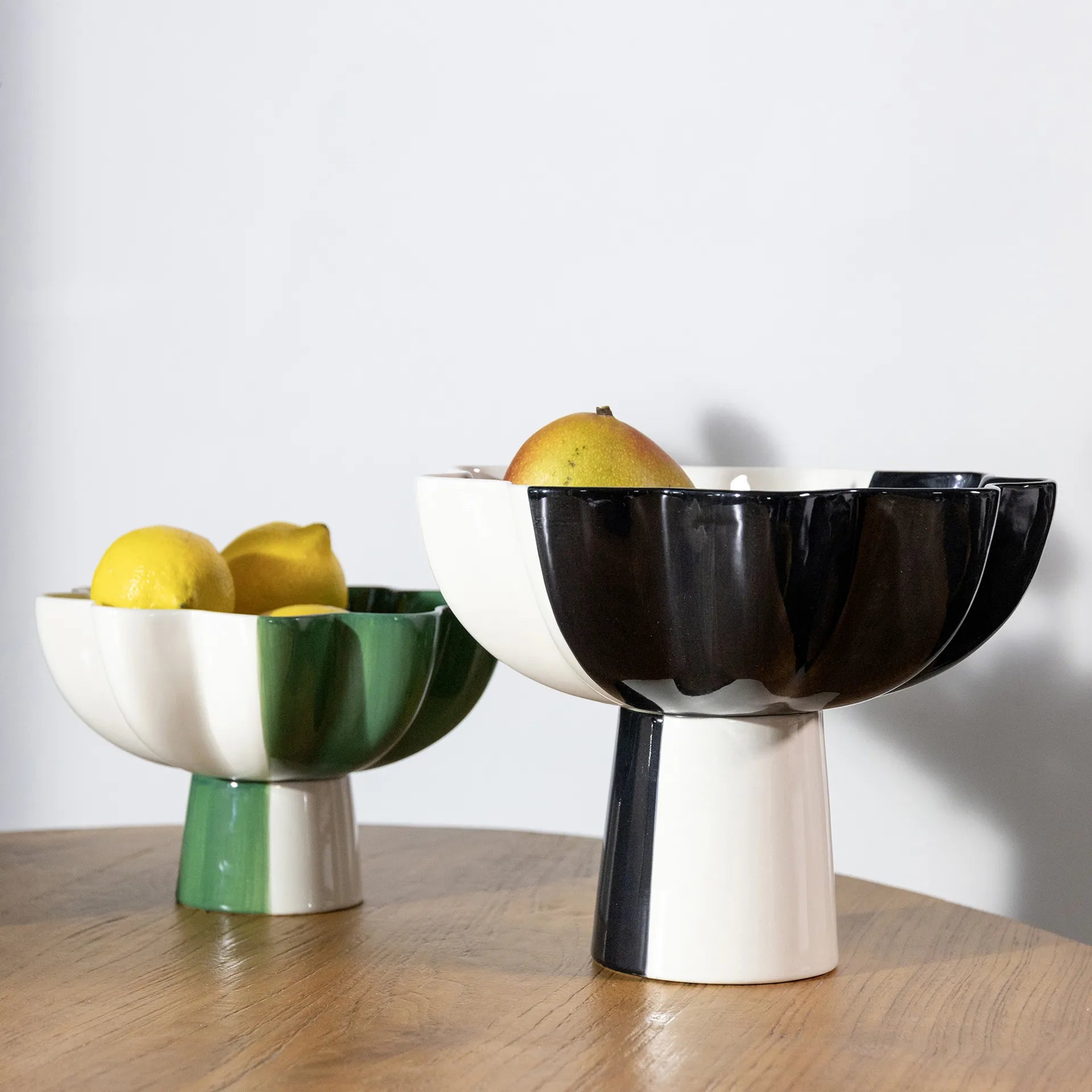 Two multicolor ceramic pedestal bowls are on a wooden surface. The left, in green and white stoneware, holds two lemons. The right bowl is the &Klevering Fruit Bowl Sun Black Large with a modern segmented design, cradling a pear and apple, adding charm to any space.