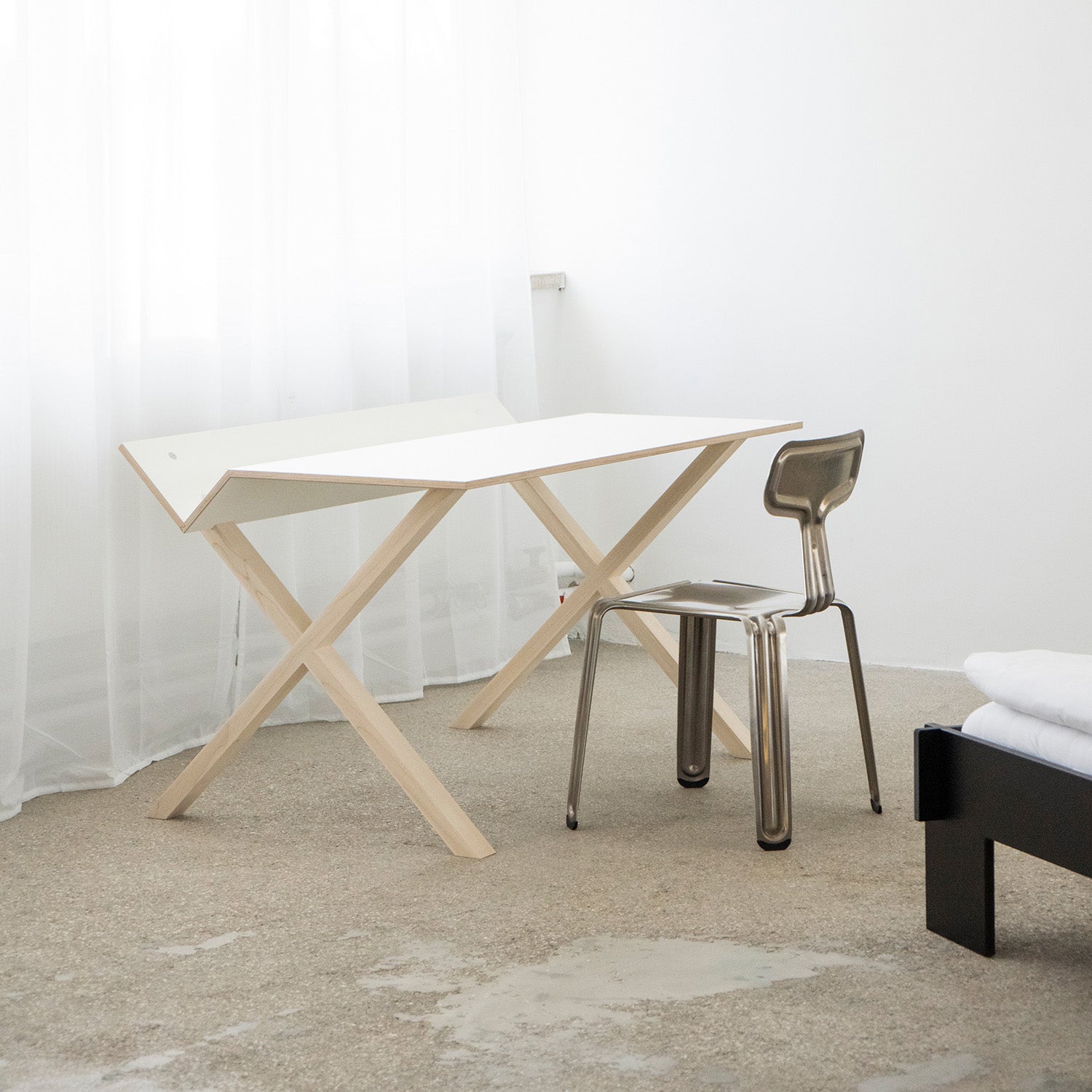 A minimalist room showcases a modern, organized desk area featuring the Kant desk by Moormann, with angular white surfaces and light wooden legs, paired with a sleek metallic chair. Sheer white curtains dress the nearby window and part of a bed with white bedding completes this clean work surface haven.