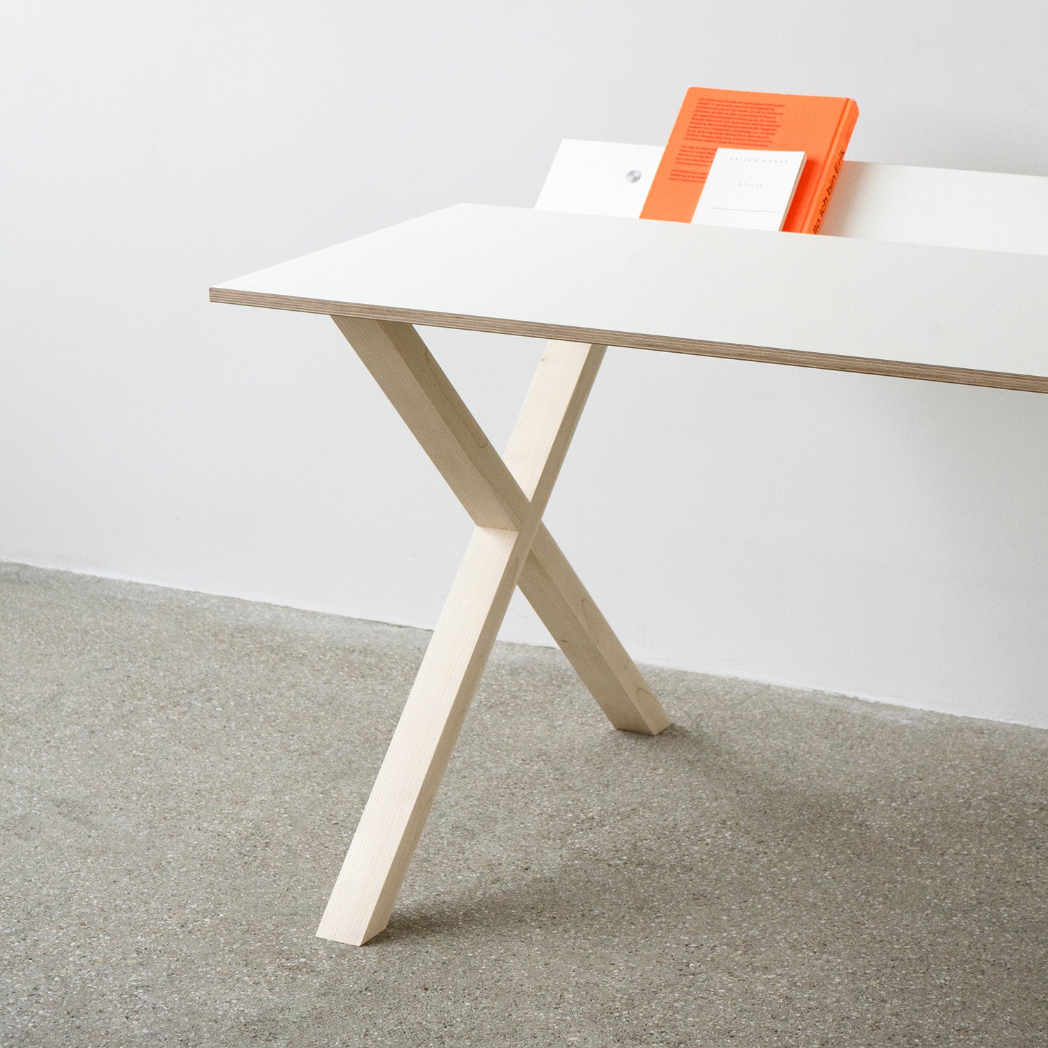 The Kant desk by Moormann, featuring an X-shaped wooden leg against a white wall, creates an organized workspace. A bright orange book rests on the pristine surface, while the light gray concrete floor enhances the tidy setup.