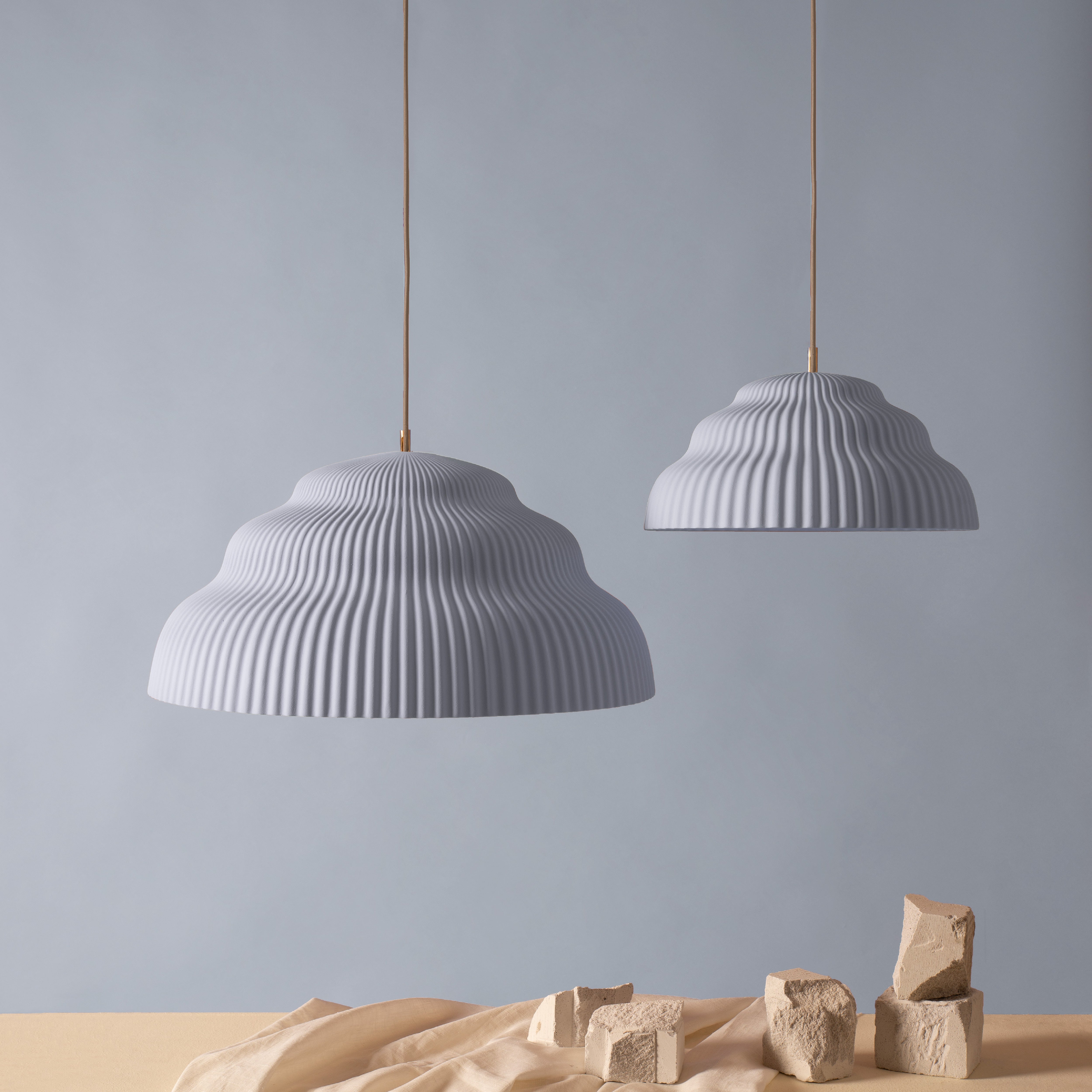 Two Kaskad Light Blue ribbed, dome-shaped pendant lamps by Schneid hang against a light gray background. Below, a beige surface is adorned with scattered clay bricks and draped fabric for a minimalist and artistic setup highlighting the elegance of Kaskad designs.