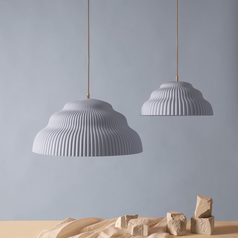 Two Kaskad Light Blue ribbed, dome-shaped pendant lamps by Schneid hang against a light gray background. Below, a beige surface is adorned with scattered clay bricks and draped fabric for a minimalist and artistic setup highlighting the elegance of Kaskad designs.