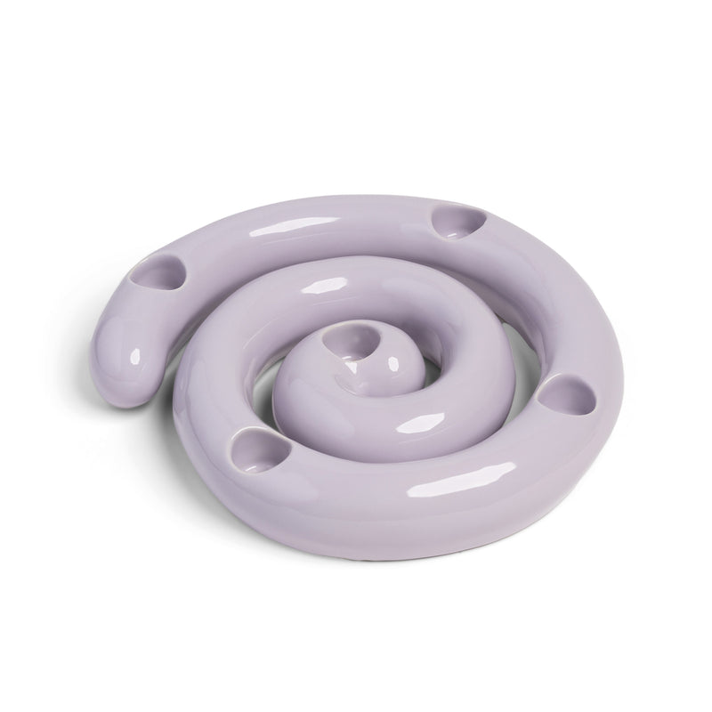 The Candle Holder Scribble by &Klevering is a glossy, lavender ceramic piece spiraling inward like a coiled snake. It resembles a porcelain candle holder with smooth, reflective curves and several evenly spaced small holes.