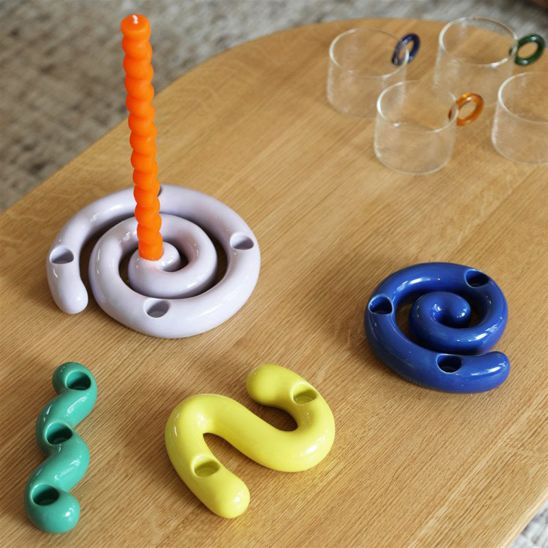&Kleverings Candle Holder Scribble designs are displayed on a wooden table, featuring colorful spiral stoneware holders in purple, blue, green, and yellow. In the background, clear glass mugs with small orange handles subtly enhance the scenes charm.