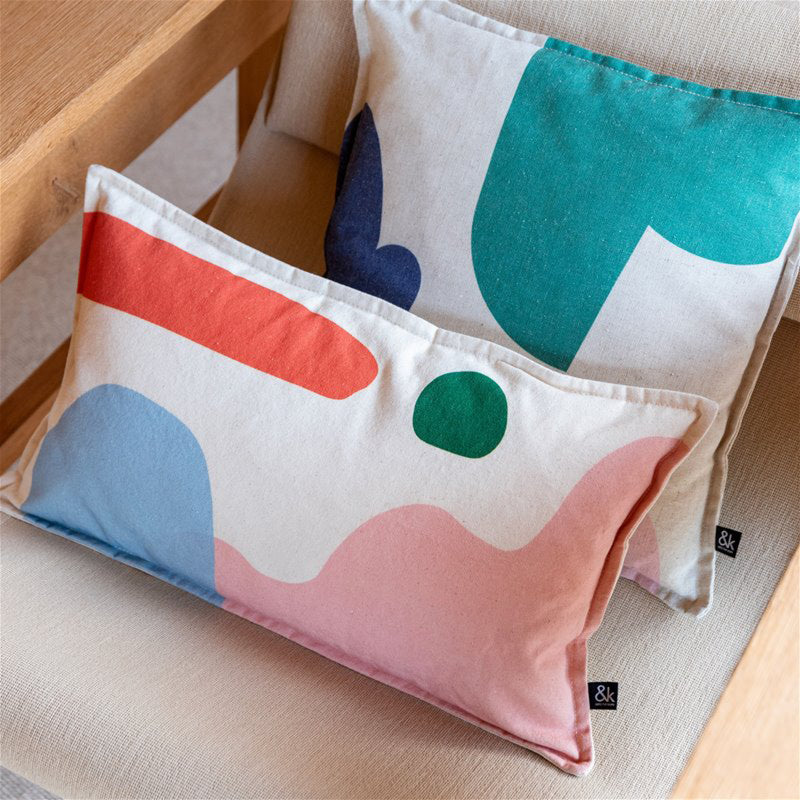 Two &Klevering Cushion Collage decorative pillows with colorful abstract shapes rest on a light-colored chair; one features red, blue, and pink designs, while the other showcases teal, green, and blue. Their high-quality cotton fabric complements the nearby wooden table.