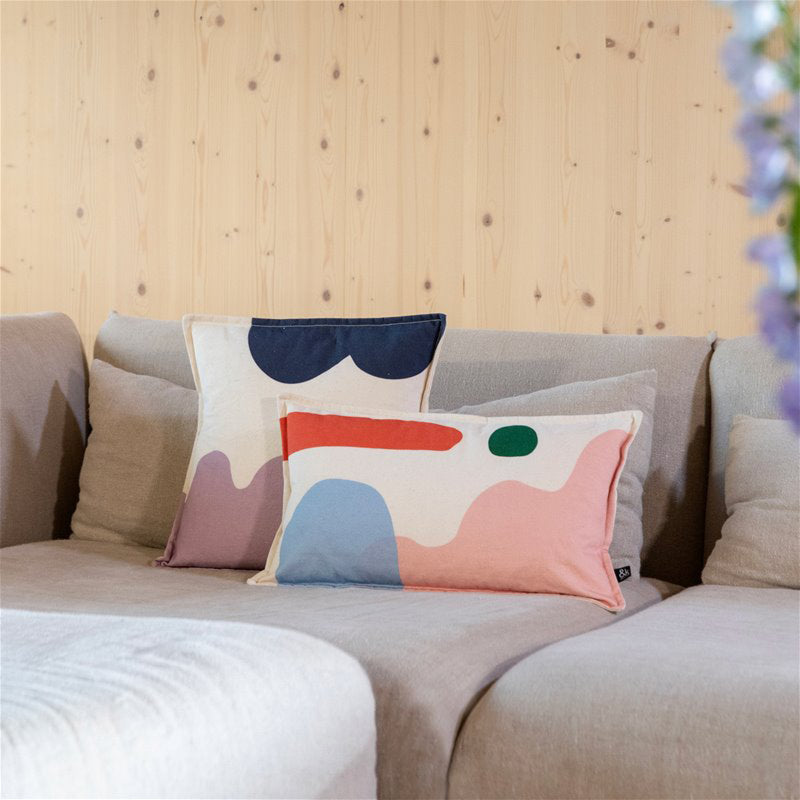 A neutral-toned sofa features the Cushion Collage by &Klevering, showcasing a high-quality cotton abstract print and a complementary decorative pillow in blue, pink, and red. The light wood-paneled wall backdrop completes this cozy and modern living space.