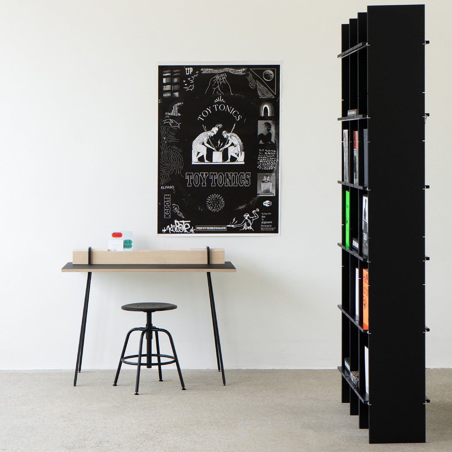 The minimalist room includes Moormanns Kurt, a simple desk paired with a black stool displaying a solid wood latch box, books, and a red toy. A black and white poster decorates the wall. On the right, Kurt also serves as a tall black bookshelf providing ample storage for colorful books on the light carpeted floor.