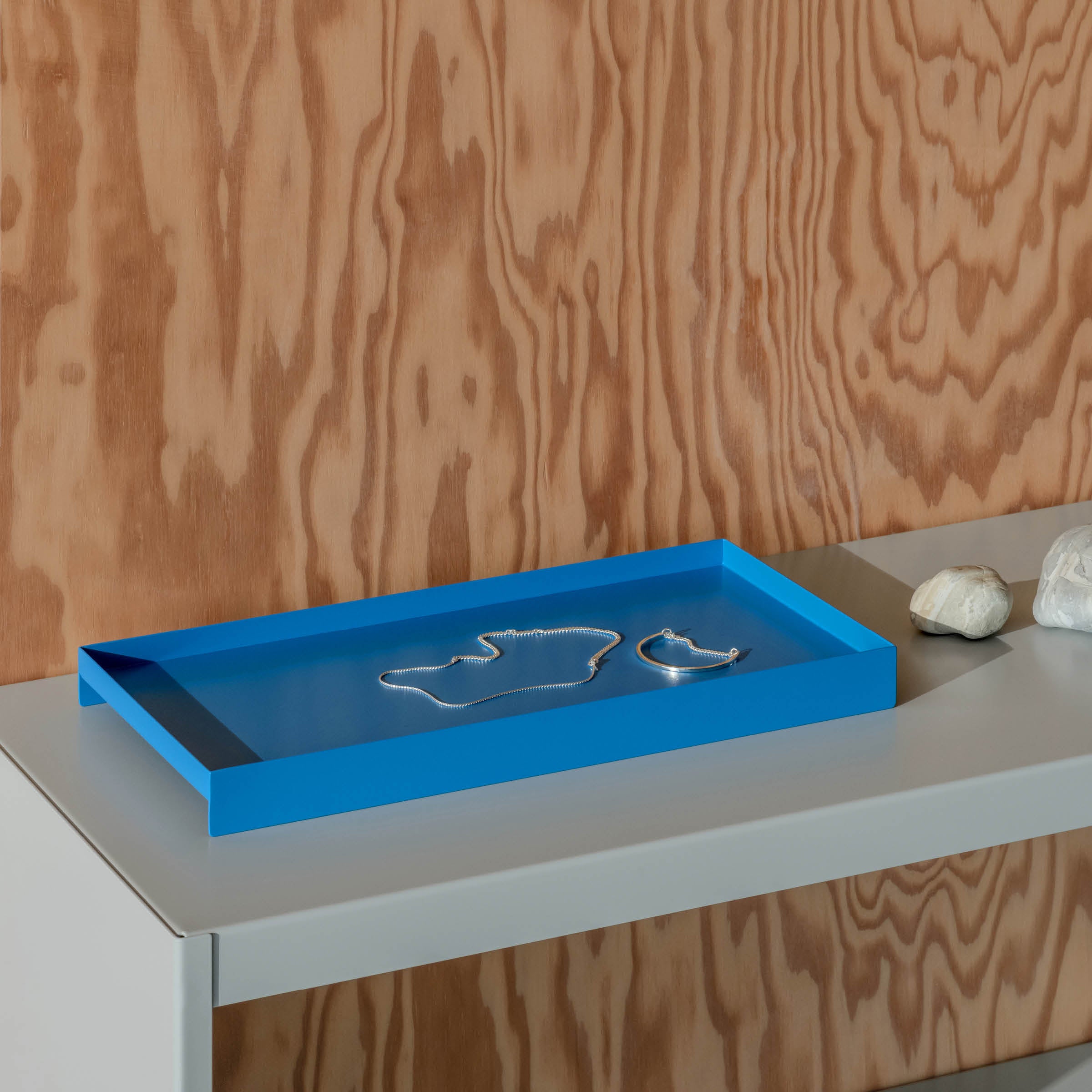 A minimalist L3 COURT steel tray by Loehr rests on a light gray table, holding two silver necklaces. The scene is set against a wooden wall with natural grain patterns. Two small white objects sit next to the tray, complementing the elegant simplicity.