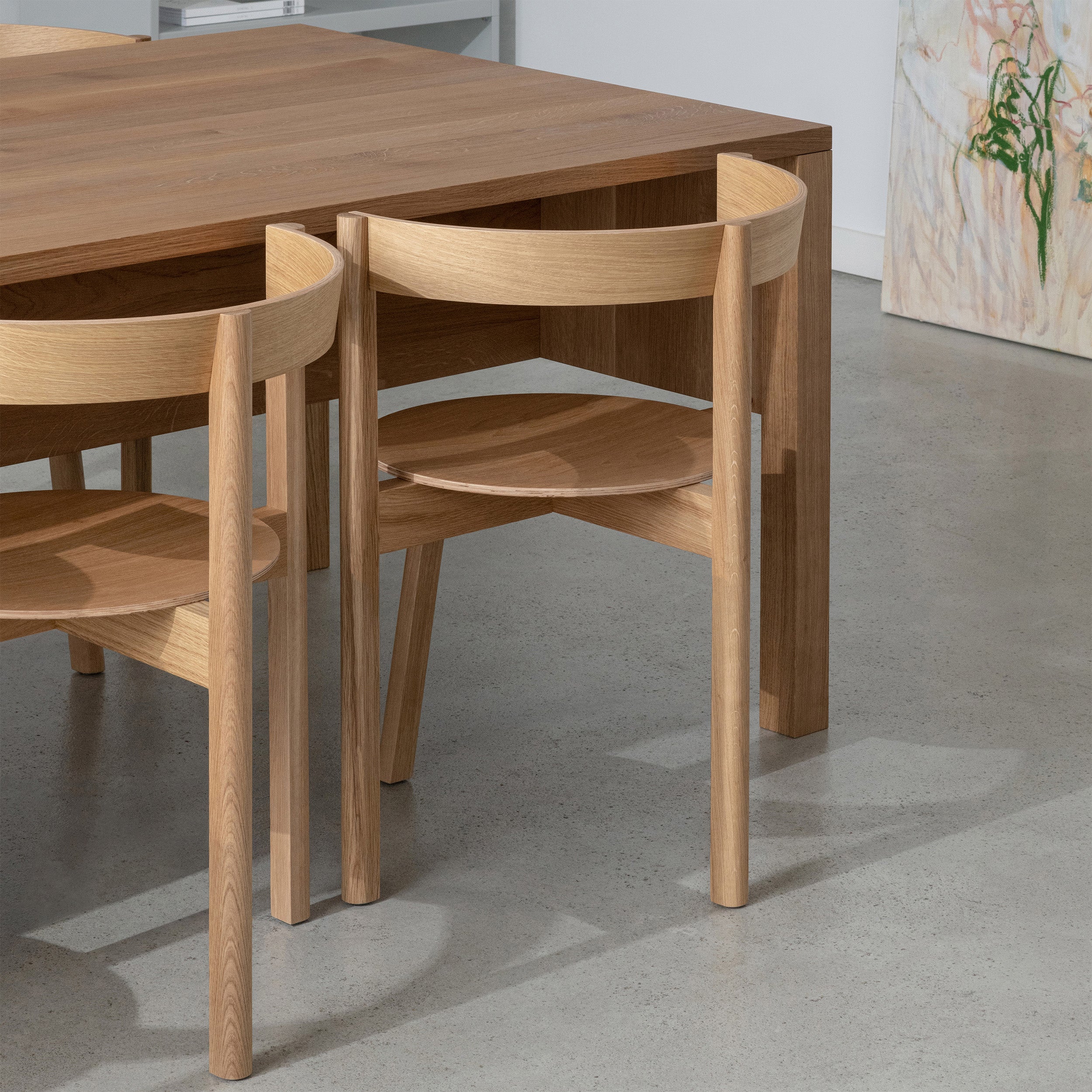 The L8 Traba Table by Loehr is complemented by wooden chairs with curved backs, all set on gray concrete flooring. The setting features various wood finishes and a piece of abstract art leaning against a white wall.