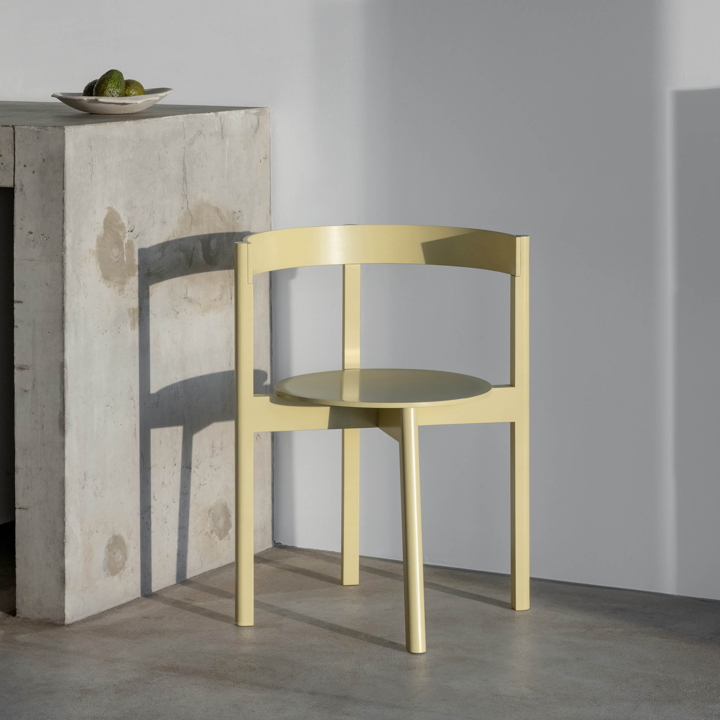 On a concrete floor, a light yellow L5 JAZZ Armchair by Loehr with a rounded backrest is paired with a concrete table. A white bowl holding green limes sits atop the table as soft natural light accents the chairs elegant design and solid wood frame.
