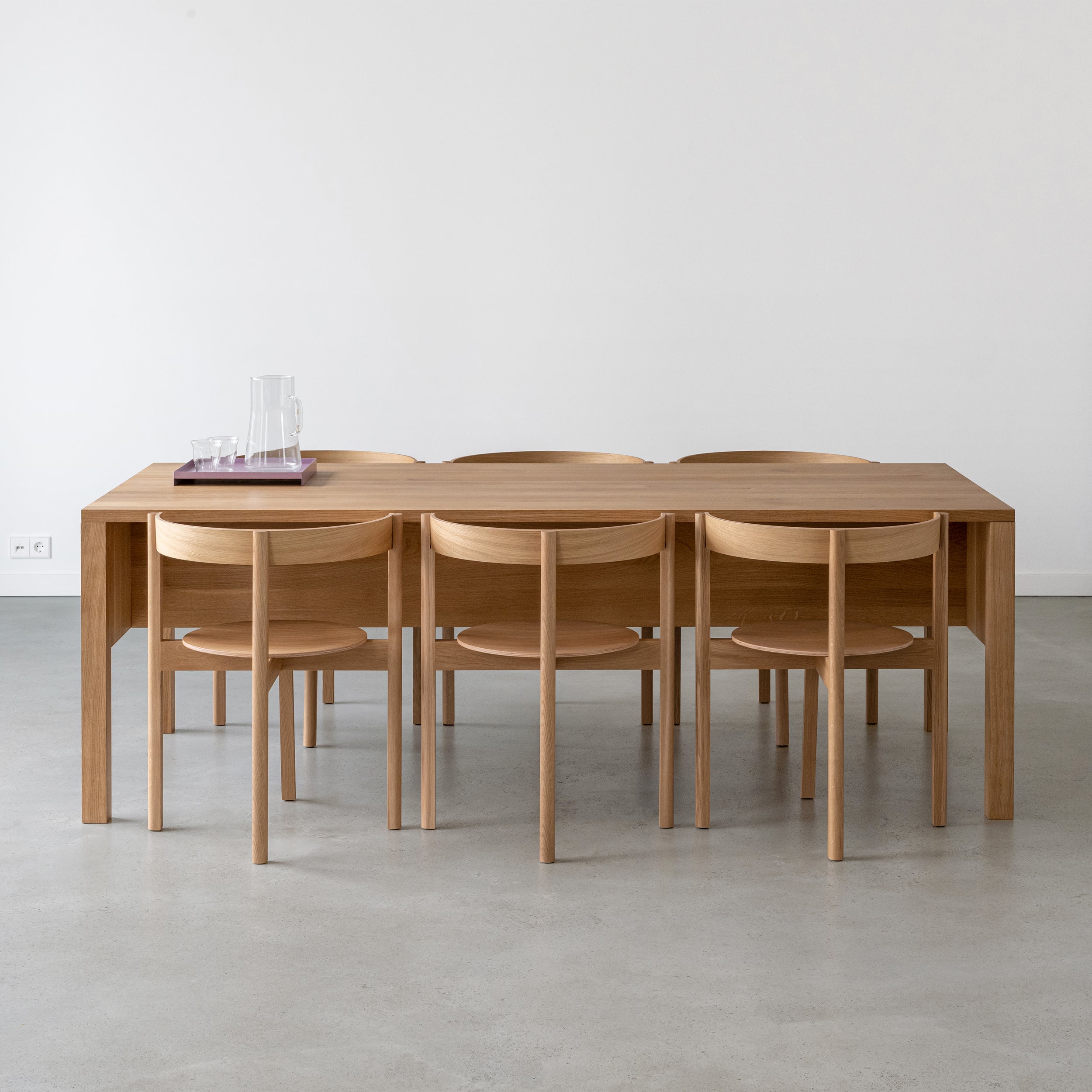 In a minimalist dining room, the L8 Traba Table by Loehr stands out, complemented by six matching chairs. A glass pitcher and four glasses on a purple tray grace the table. The serene ambiance is enhanced by diverse wood finishes, light gray floors, and white walls.